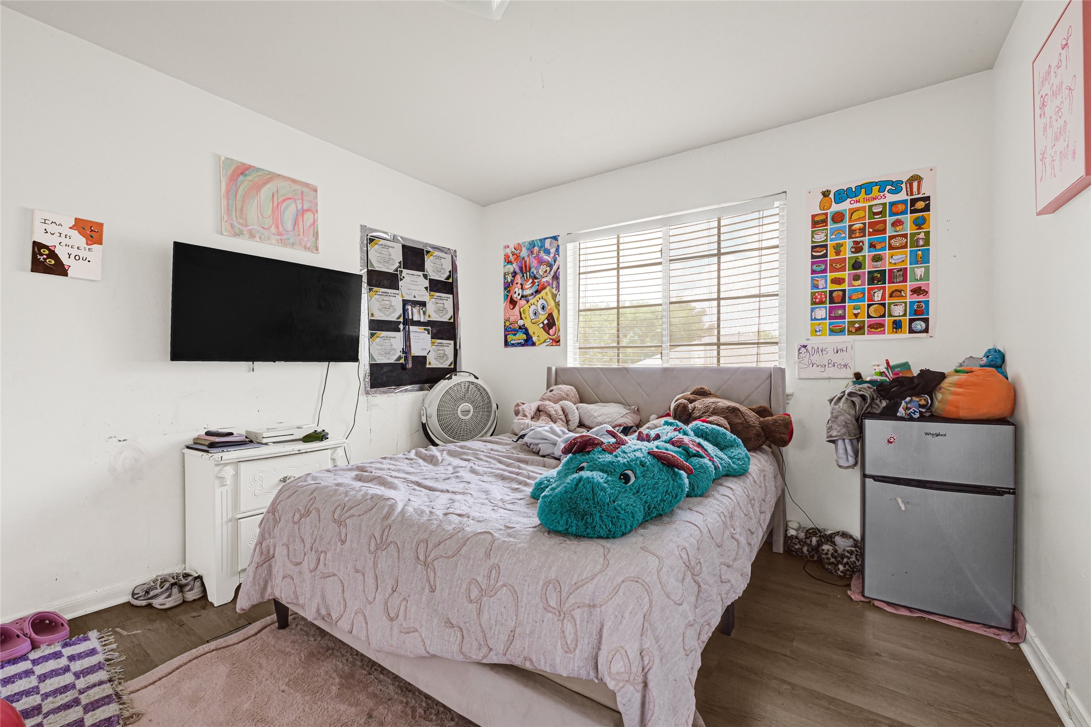 6419 Slate Stone Street Houston, TX 77084 - Photo 36 of 49 Cozy bedroom with natural light from a large window, featuring a bed adorned with plush toys. The room includes a wall-mounted TV, a small fridge, and vibrant decor, offering a warm and inviting atmosphere.