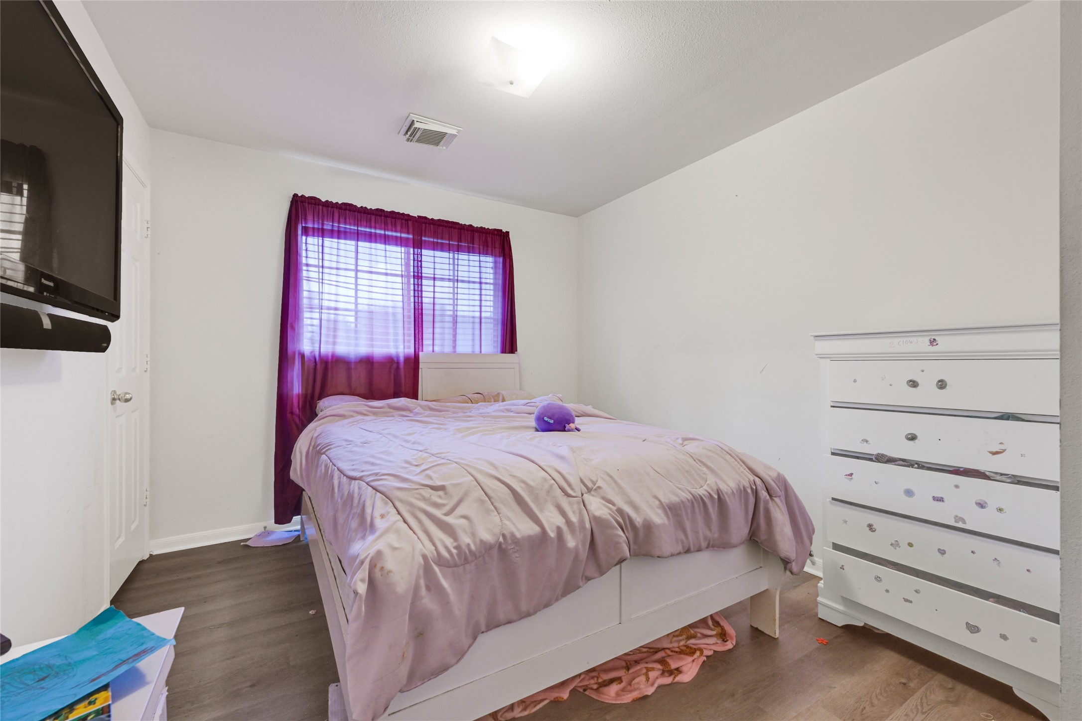 6419 Slate Stone Street Houston, TX 77084 - Photo 39 of 49 Cozy bedroom featuring a comfortable bed, wall-mounted TV, and a dresser. Natural light filters through purple curtains, complementing the neutral walls and wood flooring.