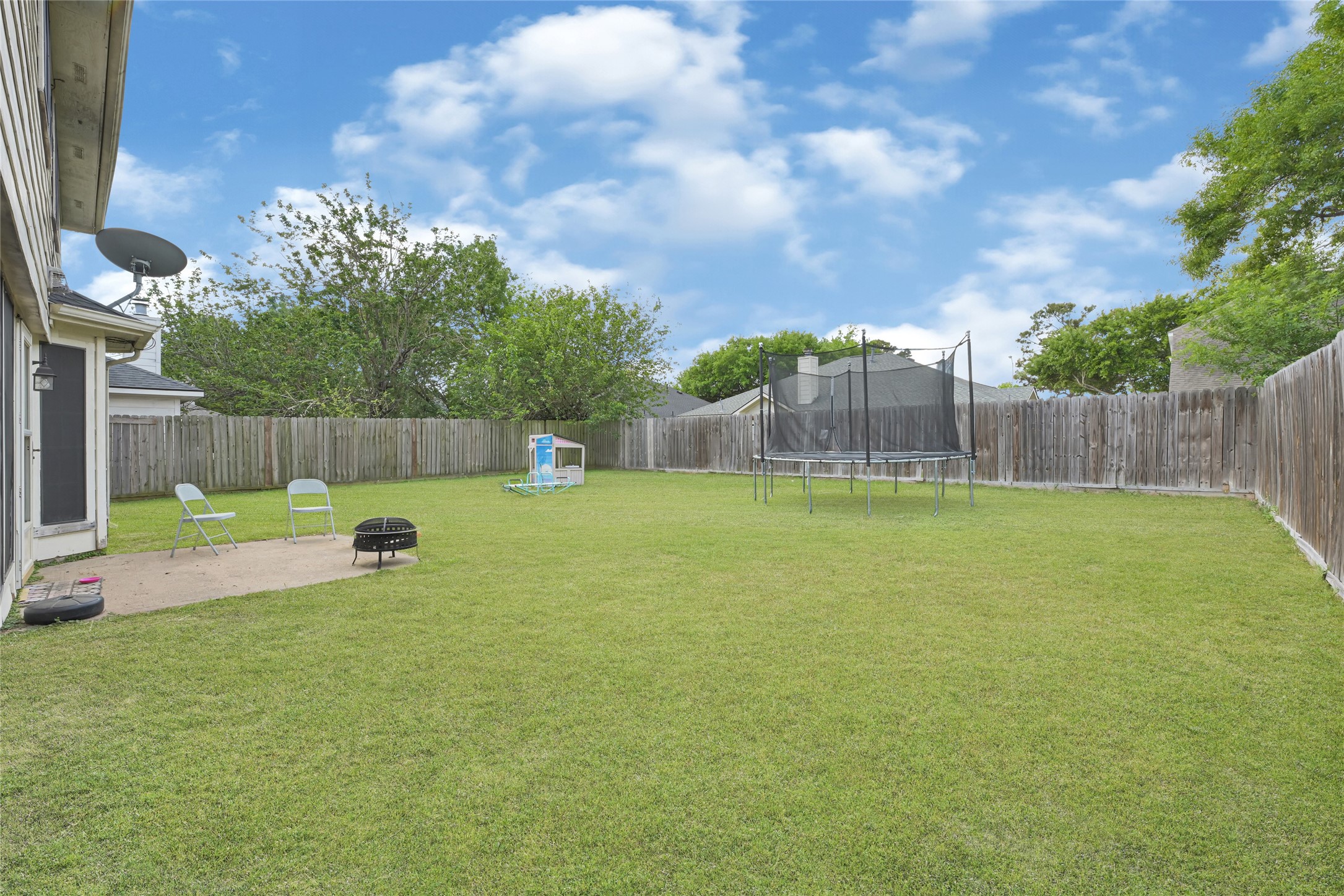 6419 Slate Stone Street Houston, TX 77084 - Photo 43 of 49 Spacious backyard with a well-maintained lawn, featuring a trampoline, patio area with chairs, and a fire pit, all enclosed by a wooden fence. Ideal for outdoor activities and relaxation.
