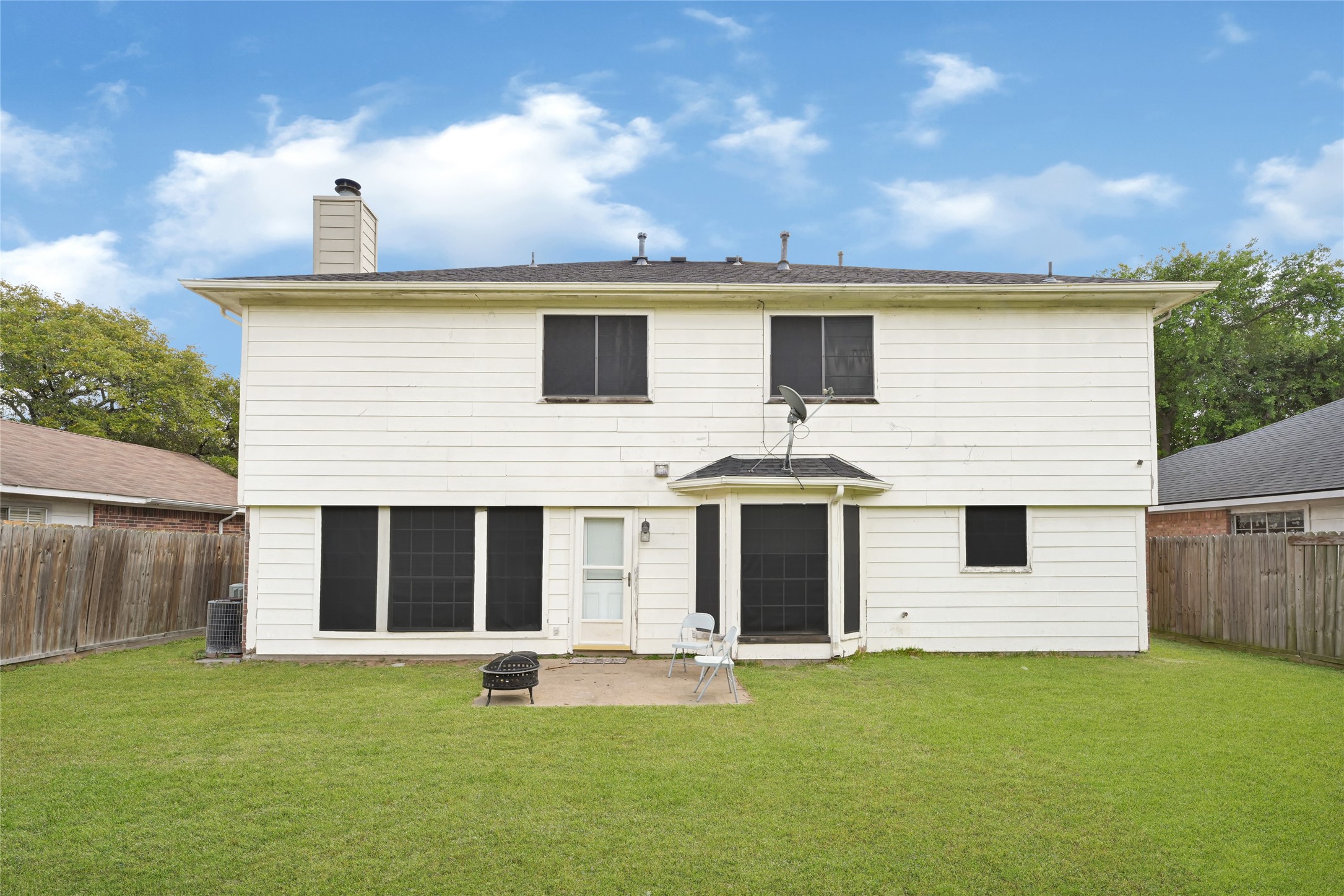6419 Slate Stone Street Houston, TX 77084 - Photo 46 of 49 This photo shows the back of a two-story home with white siding, featuring several windows and a small patio area. The yard is grassy, bordered by a wooden fence, and there is a satellite dish mounted near the back door.