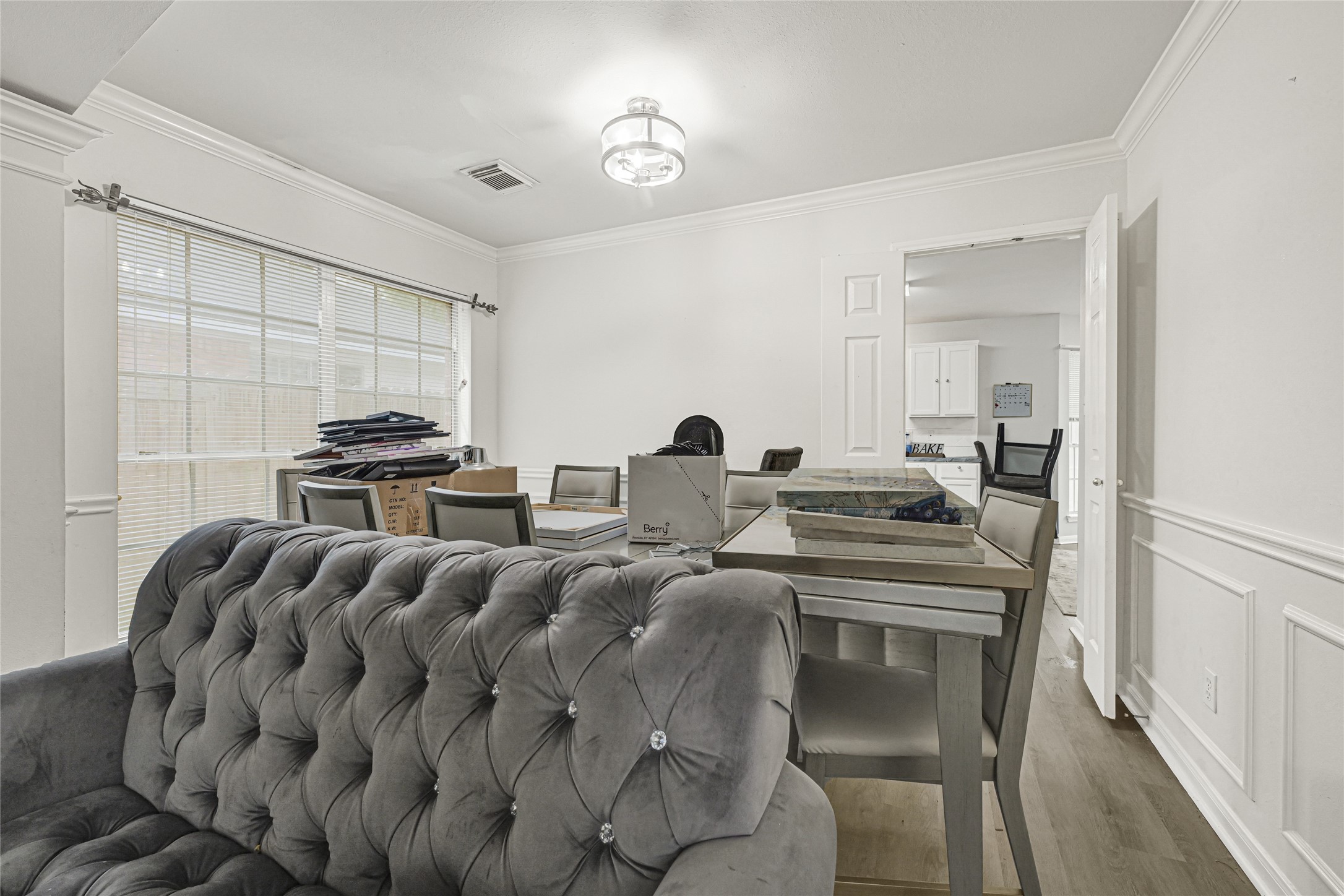 6419 Slate Stone Street Houston, TX 77084 - Photo 9 of 49 This photo showcases a bright dining space with large windows, a modern light fixture, and elegant wainscoting. The room is partially furnished with a plush tufted sofa and a dining table. Adjacent is a view into a white kitchen, enhancing the open and connected feel of the space.