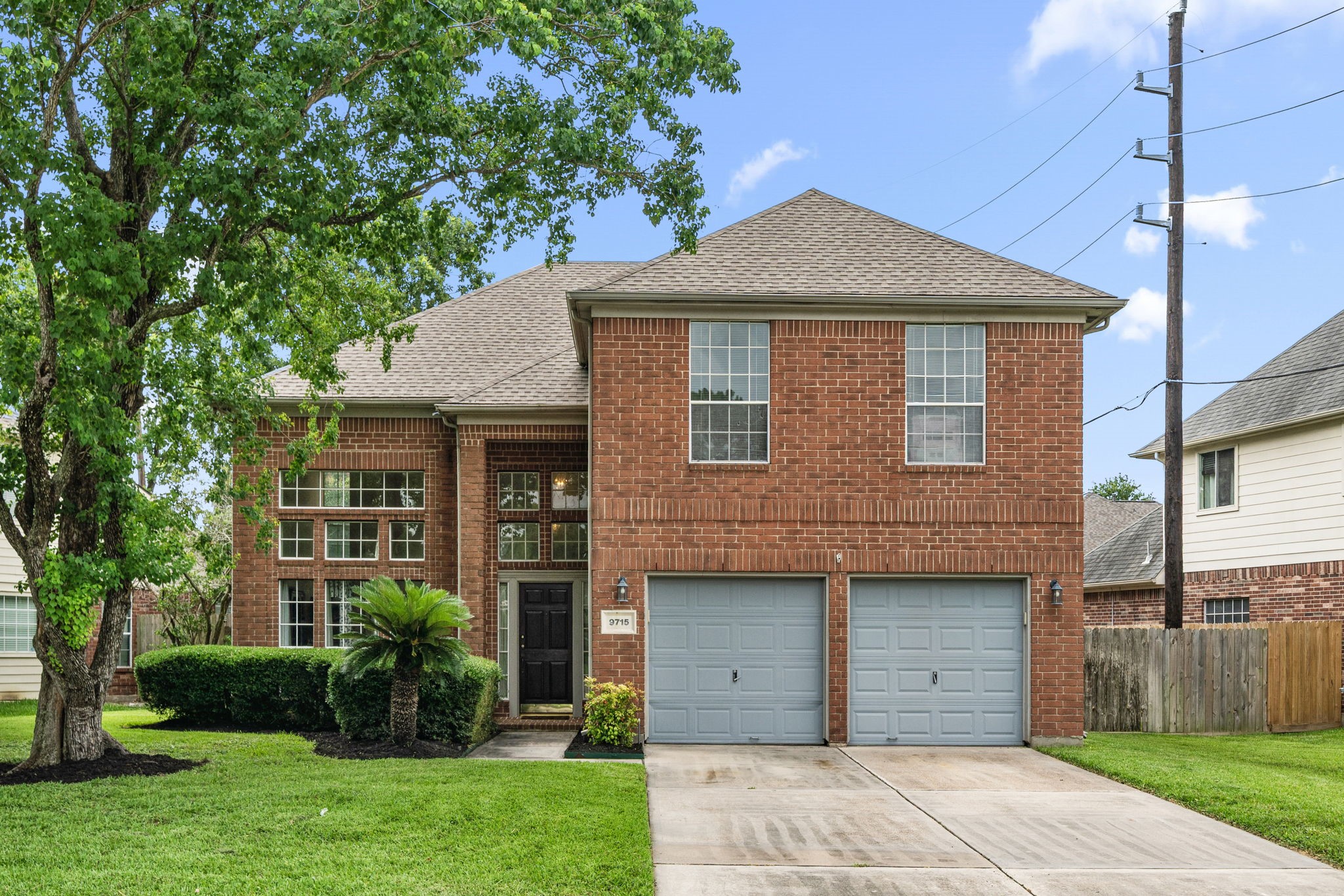This two-story brick home features a double garage, an abundance of windows, and a well-maintained front yard with mature trees.