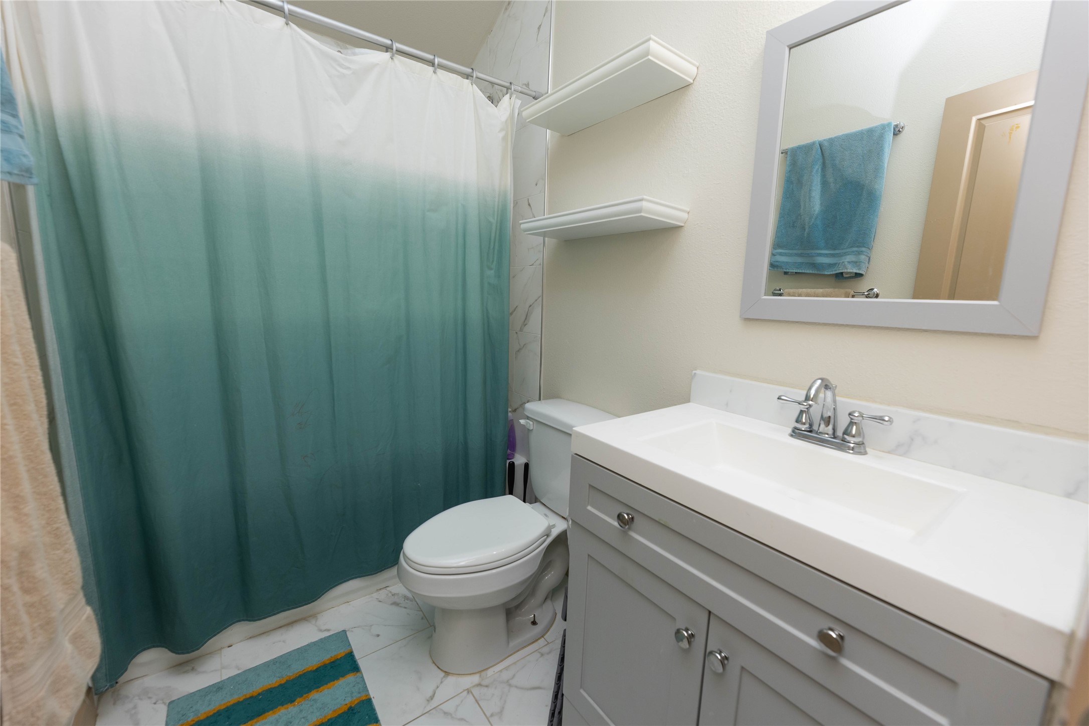 8007 Capitol Street Houston, TX 77012 - Photo 20 of 25 This bathroom features a modern design with a white vanity and marble countertop, complemented by a sleek faucet and mirror. It has a shower with a blue gradient curtain, white shelves, and a tiled floor, creating a clean and functional space.