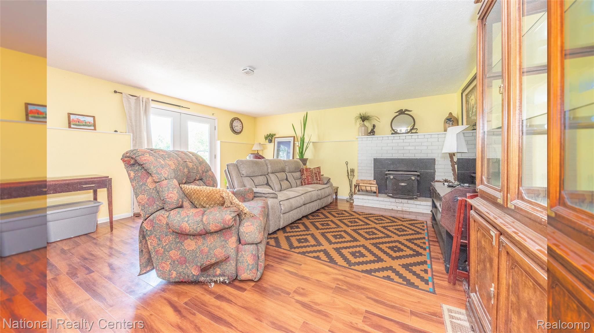 58191 8 Mile Road Northville, MI 48167 - Photo 42 of 57 Spacious living area featuring wood-finish flooring, a white brick fireplace with a dark wood stove insert, and a double patio door