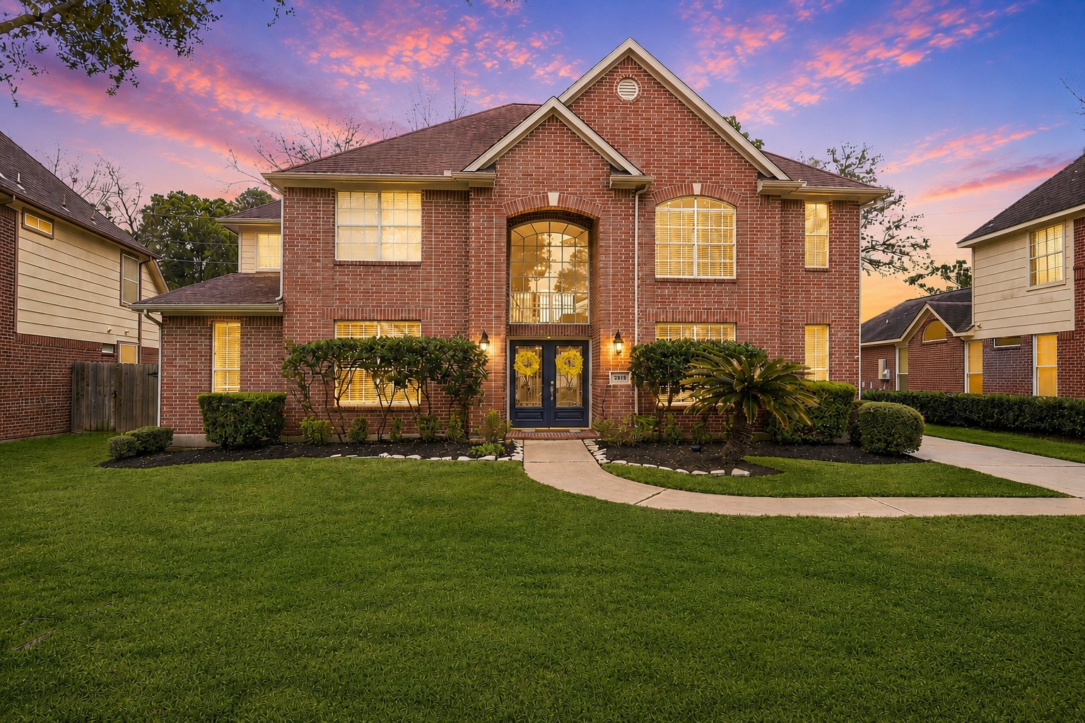 This charming two-story house with a well-maintained front yard, featuring lush green grass and landscaped shrubs. The home has a welcoming entrance with large windows and a pathway leading to the front door.