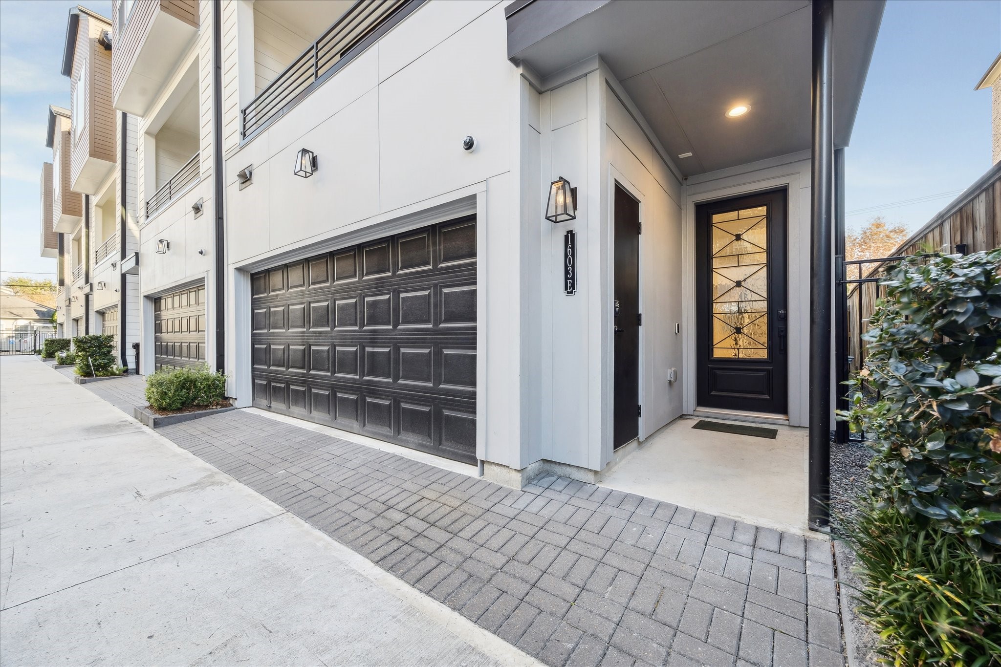 1603 Patterson Street, Unit E Houston, TX 77007 - Photo 4 of 32 This modern home features a spacious two-car garage with a sleek black door. The entrance is inviting, with a decorative glass front door and a well-lit porch. The property is part of a well-maintained row of homes, offering a clean and contemporary exterior.