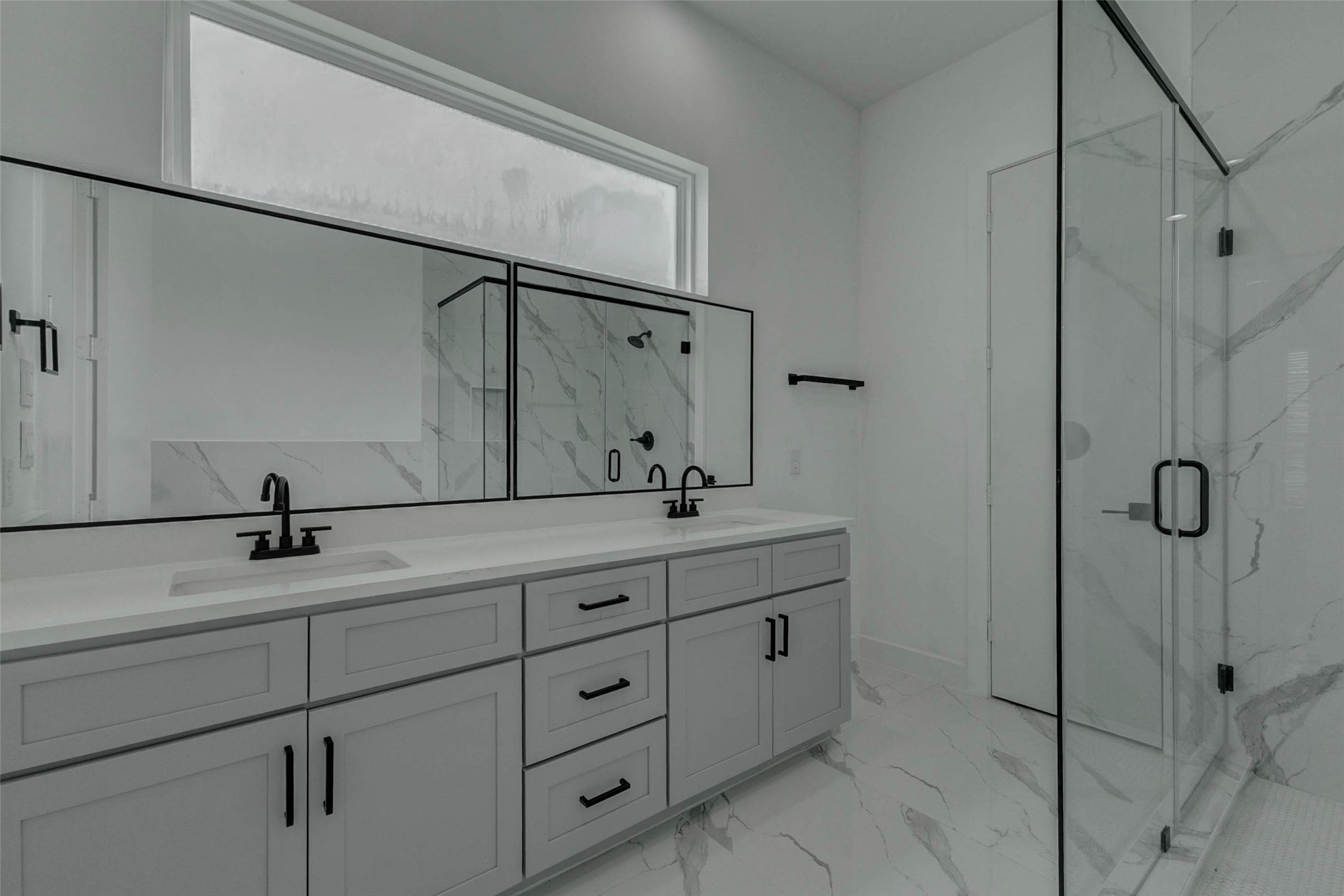 5705 Elysium Street Houston, TX 77091 - Photo 20 of 22 Primary bath with a long dual-sink vanity, soft gray cabinetry, black-framed mirror, matte black fixtures, and generous storage. The glass-enclosed shower features marble-look tile and black hardware, while the high privacy window adds natural light to the clean, spa-inspired space.