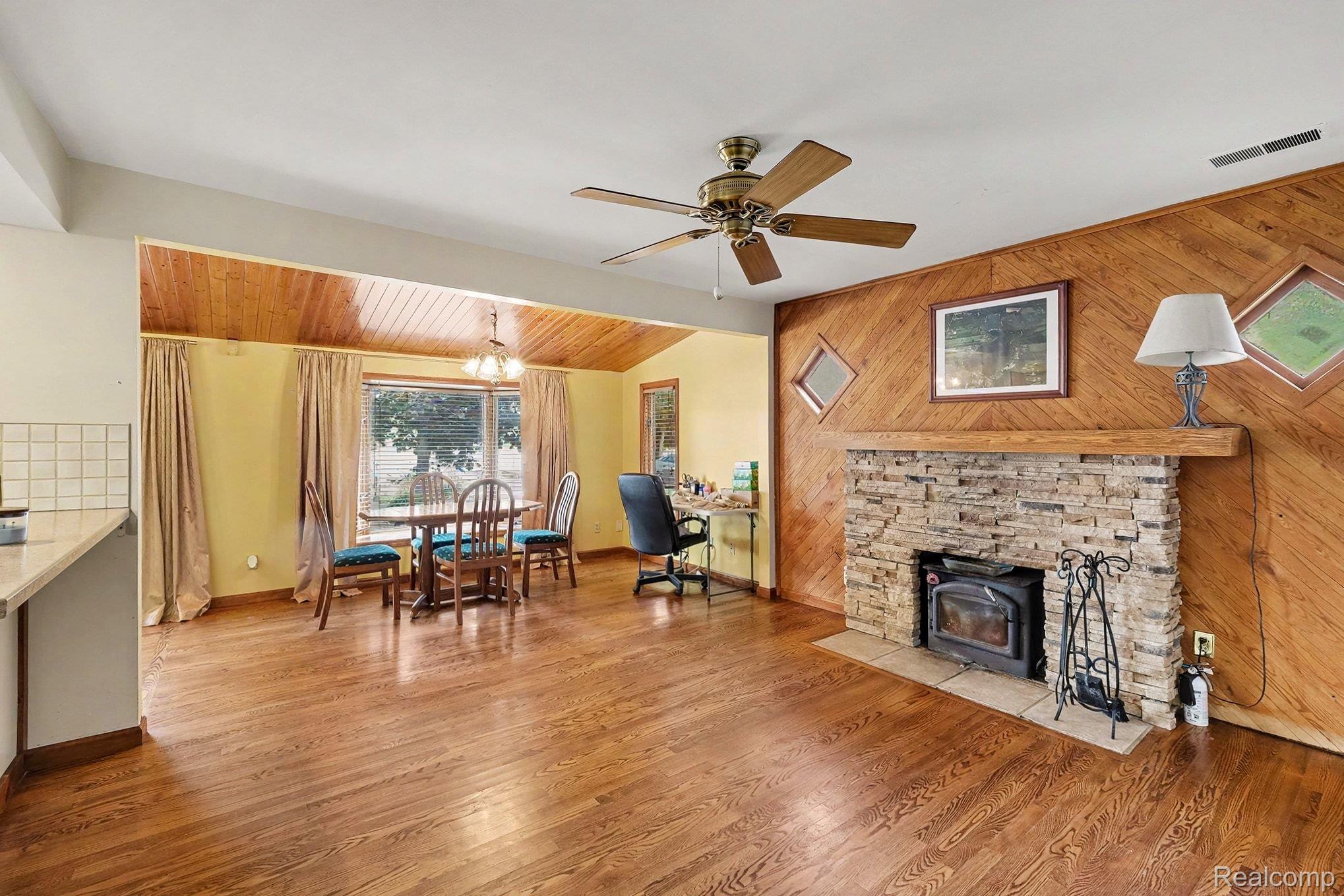 16059 Murray Road Byron, MI 48418 - Photo 7 of 32 Living area featuring wood-finish flooring, a stone-faced fireplace with a wood mantel, a wood-paneled accent wall, and a ceiling fan