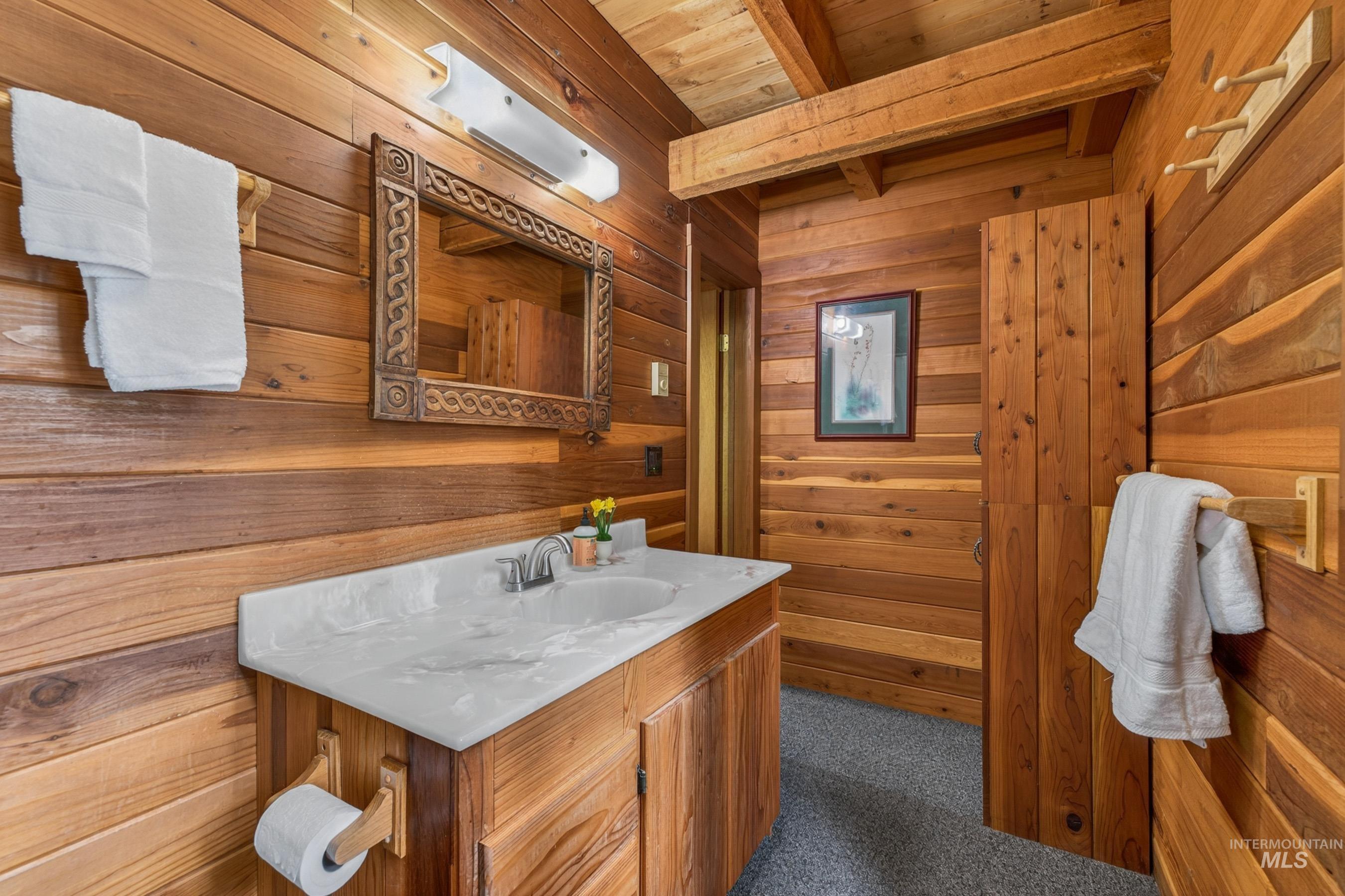 2339 West Mountain Road Donnelly, ID 83615 - Photo 37 of 37 Bathroom featuring extensive wood paneling, a wooden vanity with a single basin sink, a patterned mirror, and exposed wooden ceiling beams