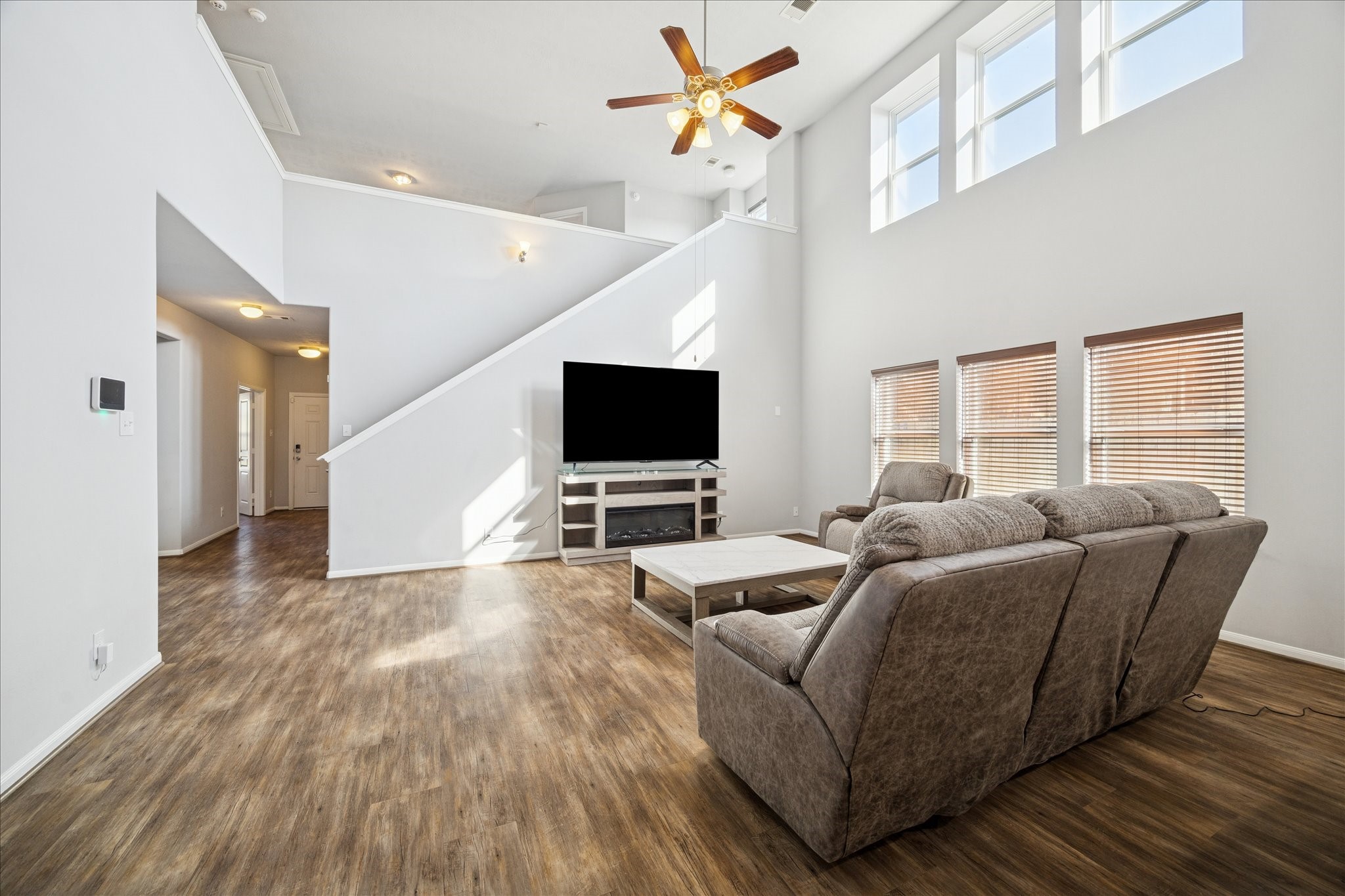 7610 Retama Terrace Lane Humble, TX 77338 - Photo 2 of 14 Spacious and bright living room featuring high ceilings, large windows, and wood flooring. It includes a cozy seating area, a ceiling fan, and a modern staircase leading to an upper level, creating an inviting and open atmosphere.