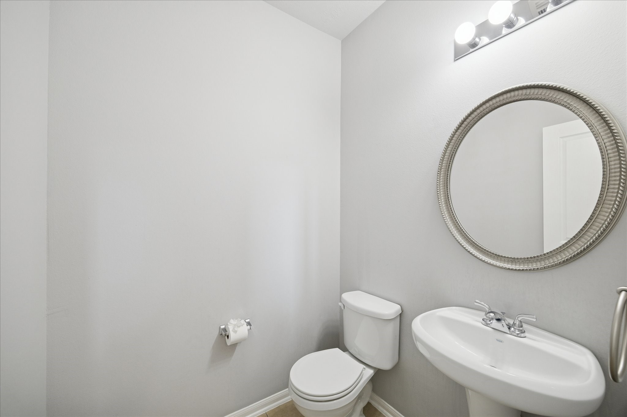 7610 Retama Terrace Lane Humble, TX 77338 - Photo 5 of 14 This bathroom features a modern, minimalist design with a pedestal sink, a round mirror, and a toilet. The space is well-lit with a sleek light fixture and has neutral wall colors for a clean, spacious feel.