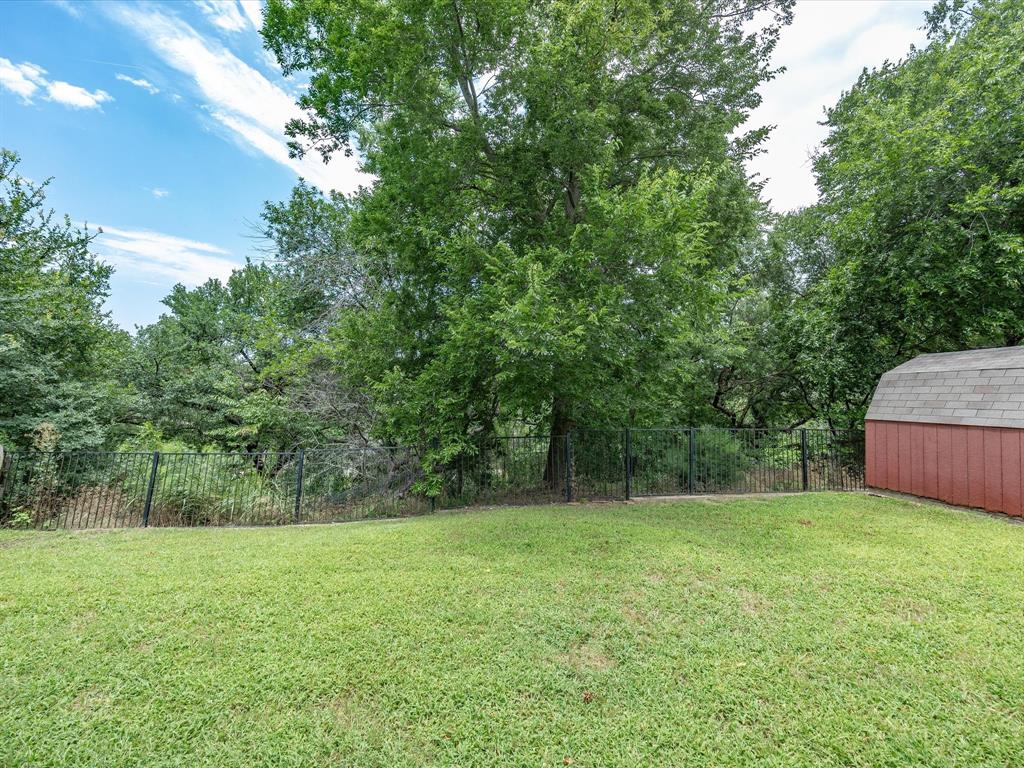 4008 Falcon Ridge Drive Haltom City, TX 76137 - Photo 39 of 40 Expansive grassy yard area featuring a black metal fence, mature trees, and a red storage shed with a shingled roof