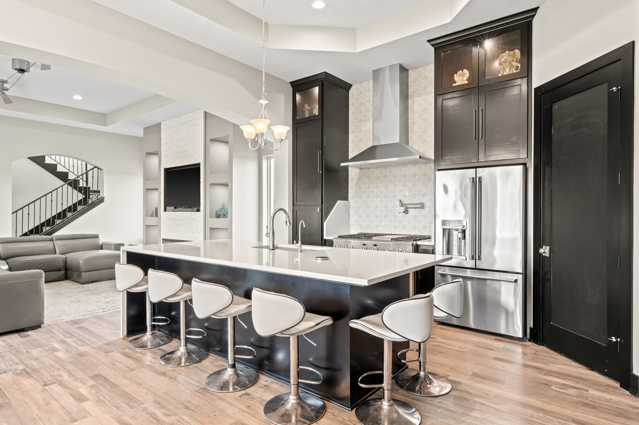 2856 Wolcek Road Crosby, TX 77532 - Photo 12 of 48 This modern "smart" kitchen features a spacious island with seating, stainless steel appliances, and sleek soft-close cabinetry doors. It opens up to a cozy living area with a sectional sofa and built-in shelving, highlighted by a beautifully tiled staircase in the background.