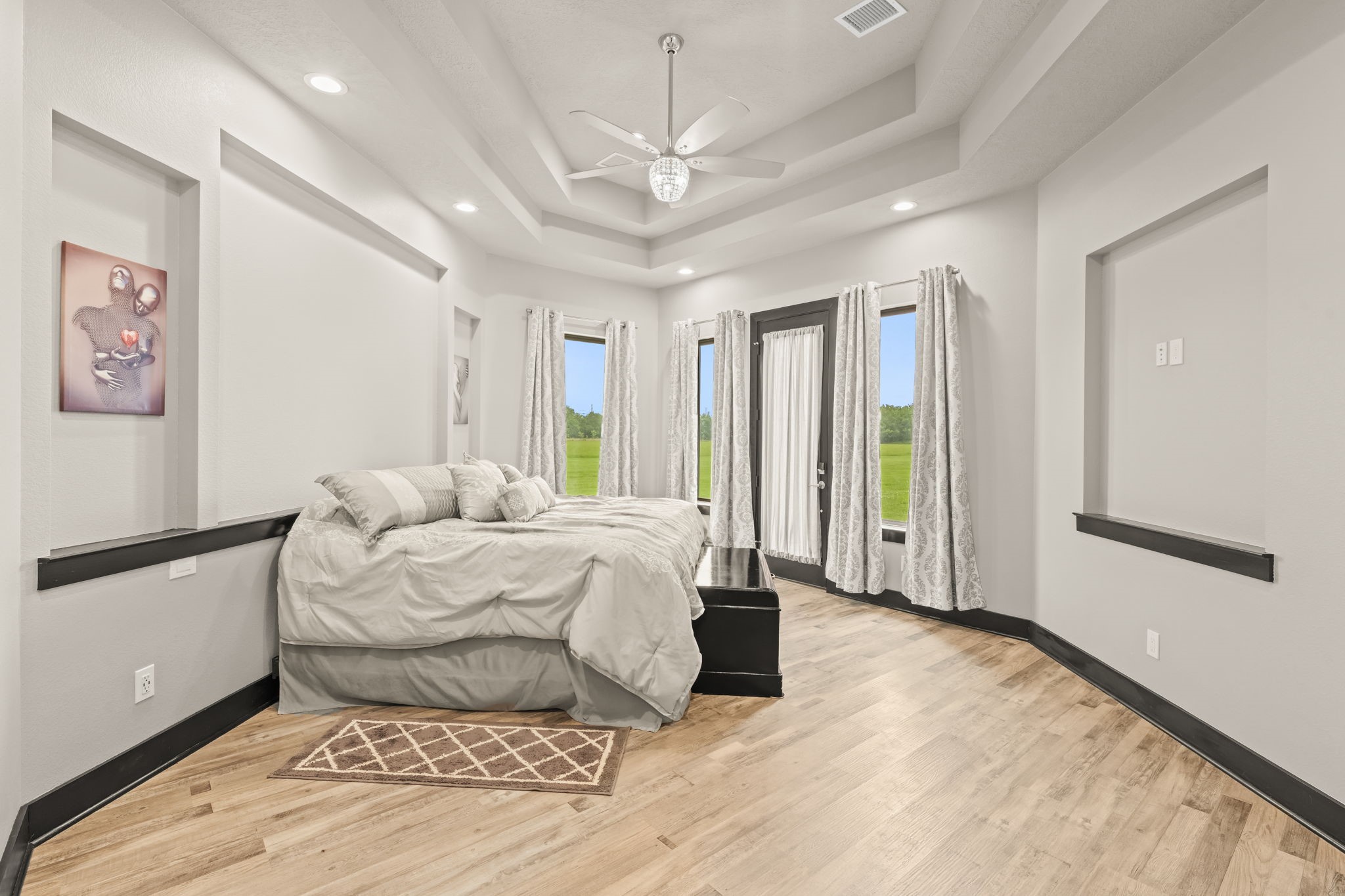 2856 Wolcek Road Crosby, TX 77532 - Photo 18 of 48 This spacious bedroom features a modern design with tray ceilings and recessed lighting. Large windows and a glass door provide abundant natural light and scenic views. The room is styled in neutral tones with light engineered wood flooring, offering a serene and inviting atmosphere.