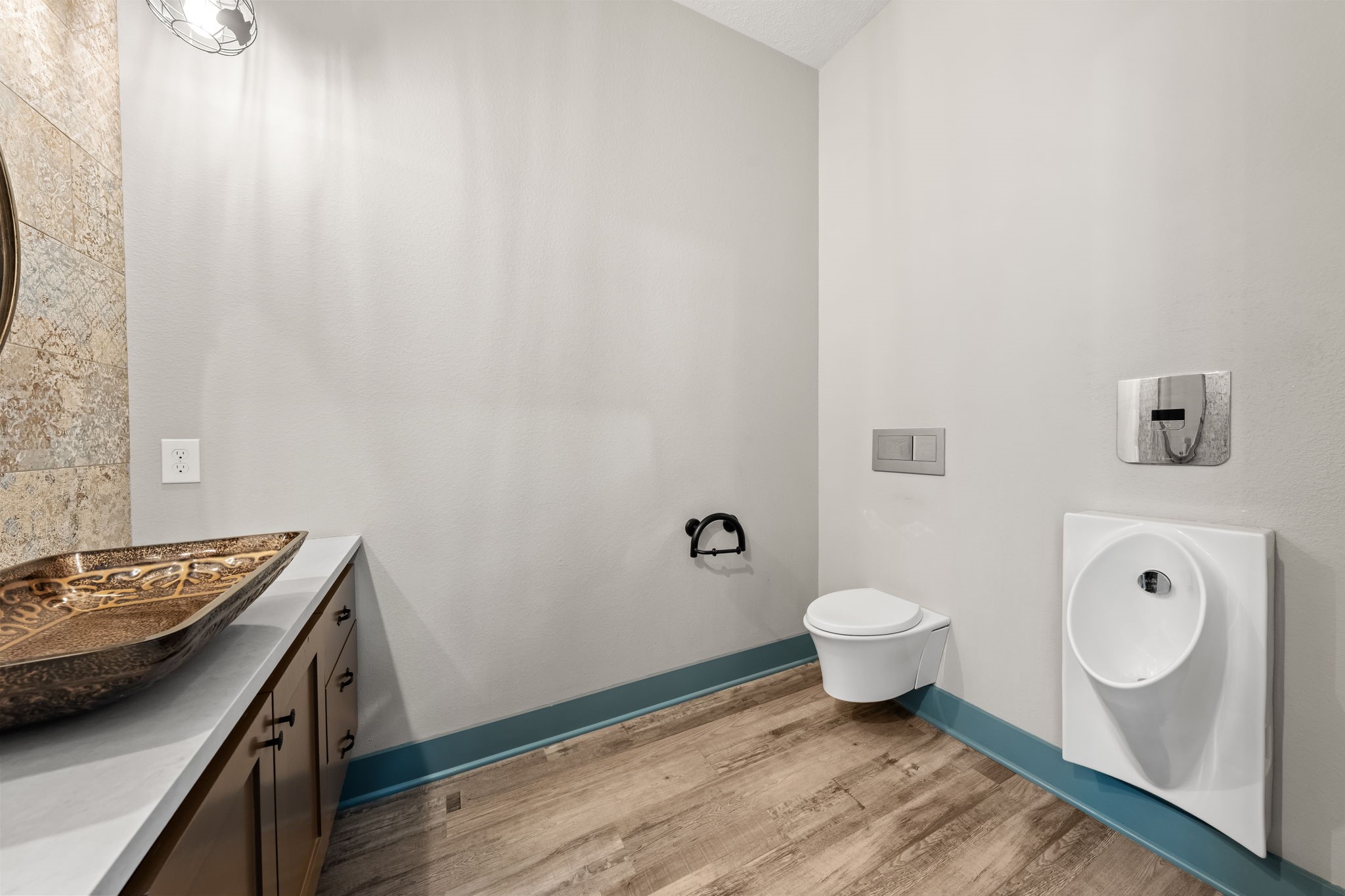2856 Wolcek Road Crosby, TX 77532 - Photo 25 of 48 This modern half bathroom features a wall-mounted toilet and urinal, a stylish vessel sink on a sleek countertop, and wood-like flooring, offering a contemporary and functional design.