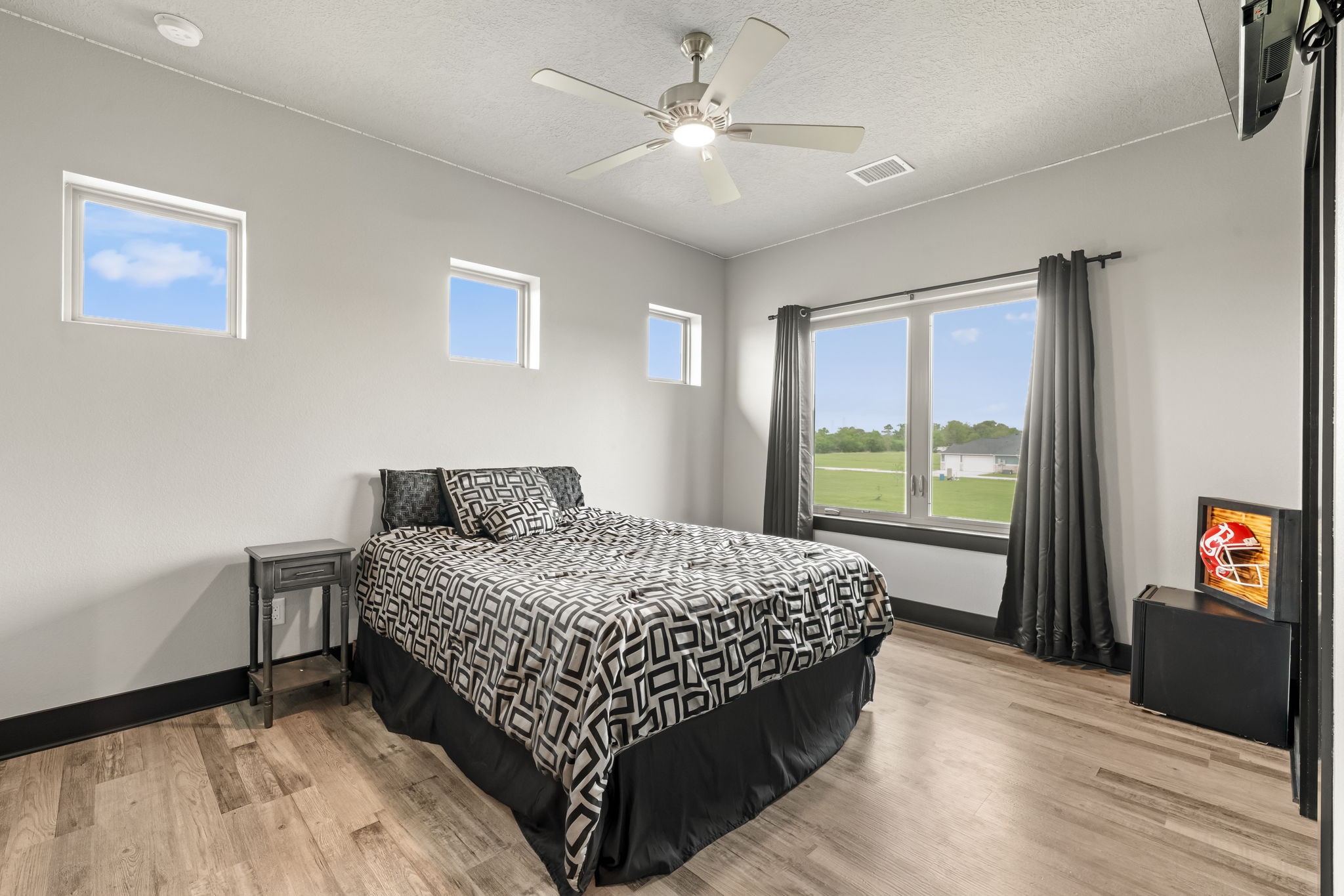 2856 Wolcek Road Crosby, TX 77532 - Photo 30 of 48 This upstairs ensuite bedroom features a modern design with light gray walls and wood flooring. It has three small windows for natural light, and a larger window with curtains offering a scenic view. The ceiling fan adds comfort and style.