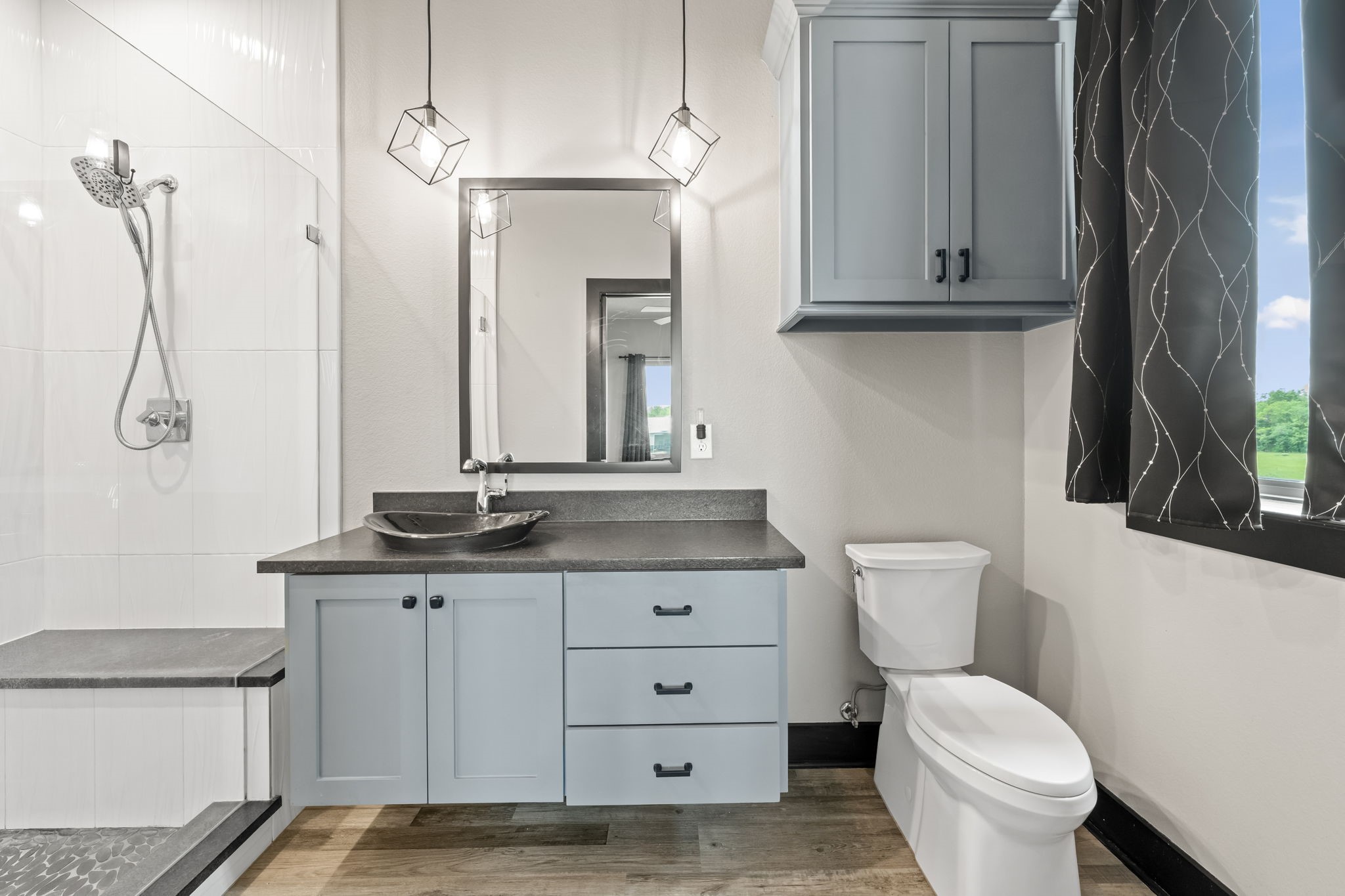 2856 Wolcek Road Crosby, TX 77532 - Photo 32 of 48 This modern bathroom features a sleek design with a glass-enclosed shower, stylish gray cabinetry, and a contemporary sink. Natural light streams in through a large window, complementing the elegant fixtures and wood-look flooring.