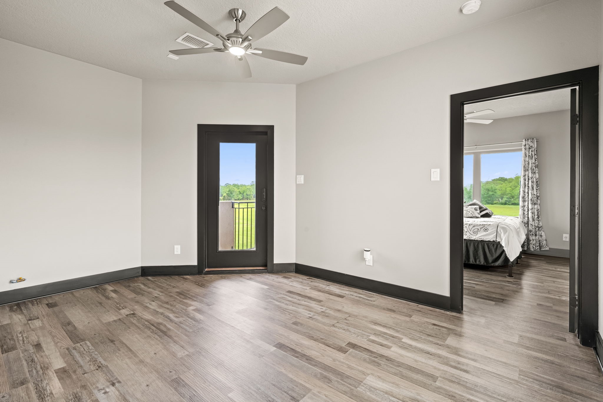 2856 Wolcek Road Crosby, TX 77532 - Photo 33 of 48 The upstairs game room with modern gray walls and engineered wood flooring. It features a ceiling fan and a door leading to a small balcony with a view of greenery. An adjacent ensuite bedroom with a large window and curtains is visible, suggesting a spacious, airy layout.