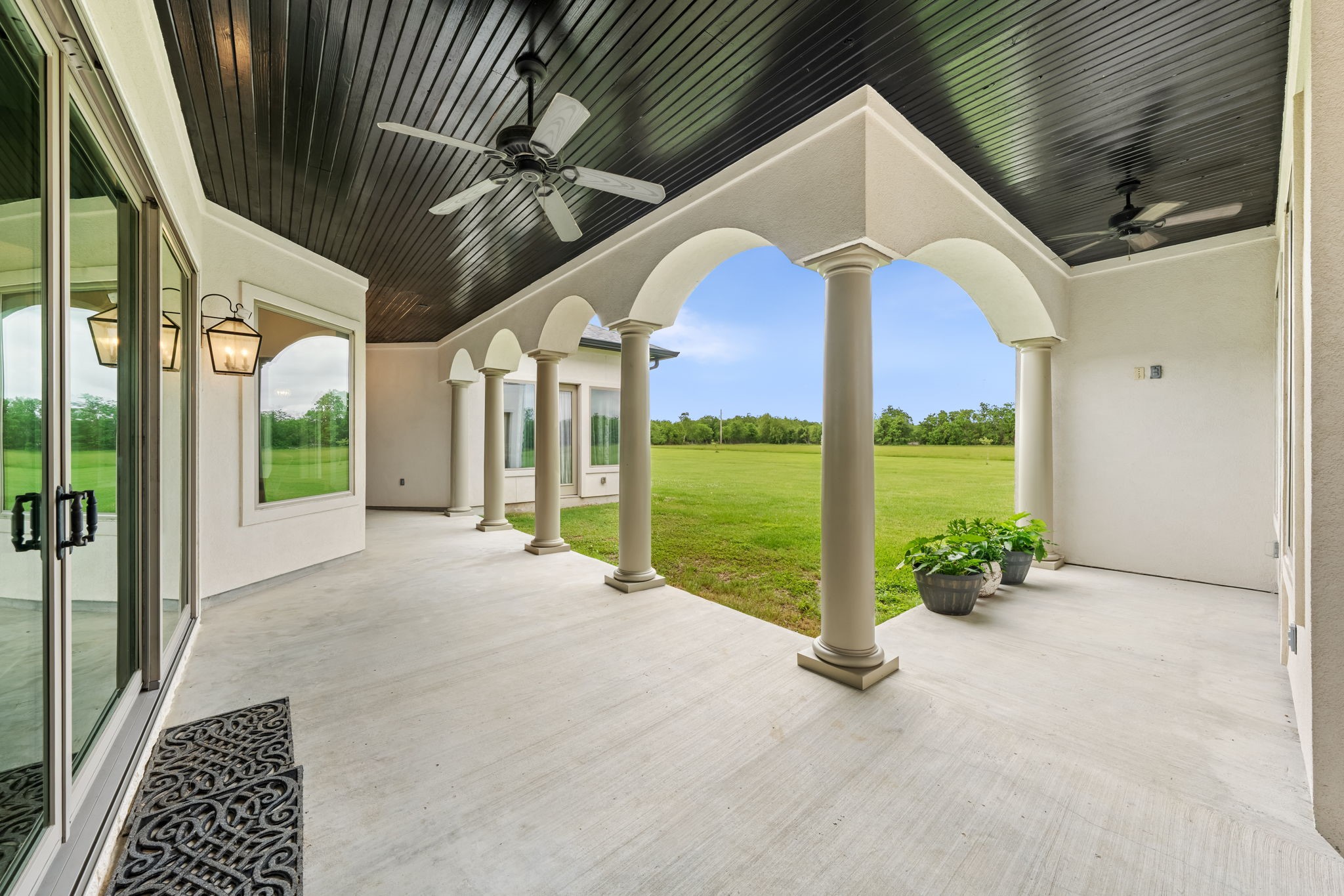 2856 Wolcek Road Crosby, TX 77532 - Photo 7 of 48 This photo showcases a spacious, elegant patio with arched columns and a dark wood ceiling featuring ceiling fans. Large glass doors provide access, and the area overlooks a lush, expansive lawn, perfect for outdoor relaxation and entertaining.