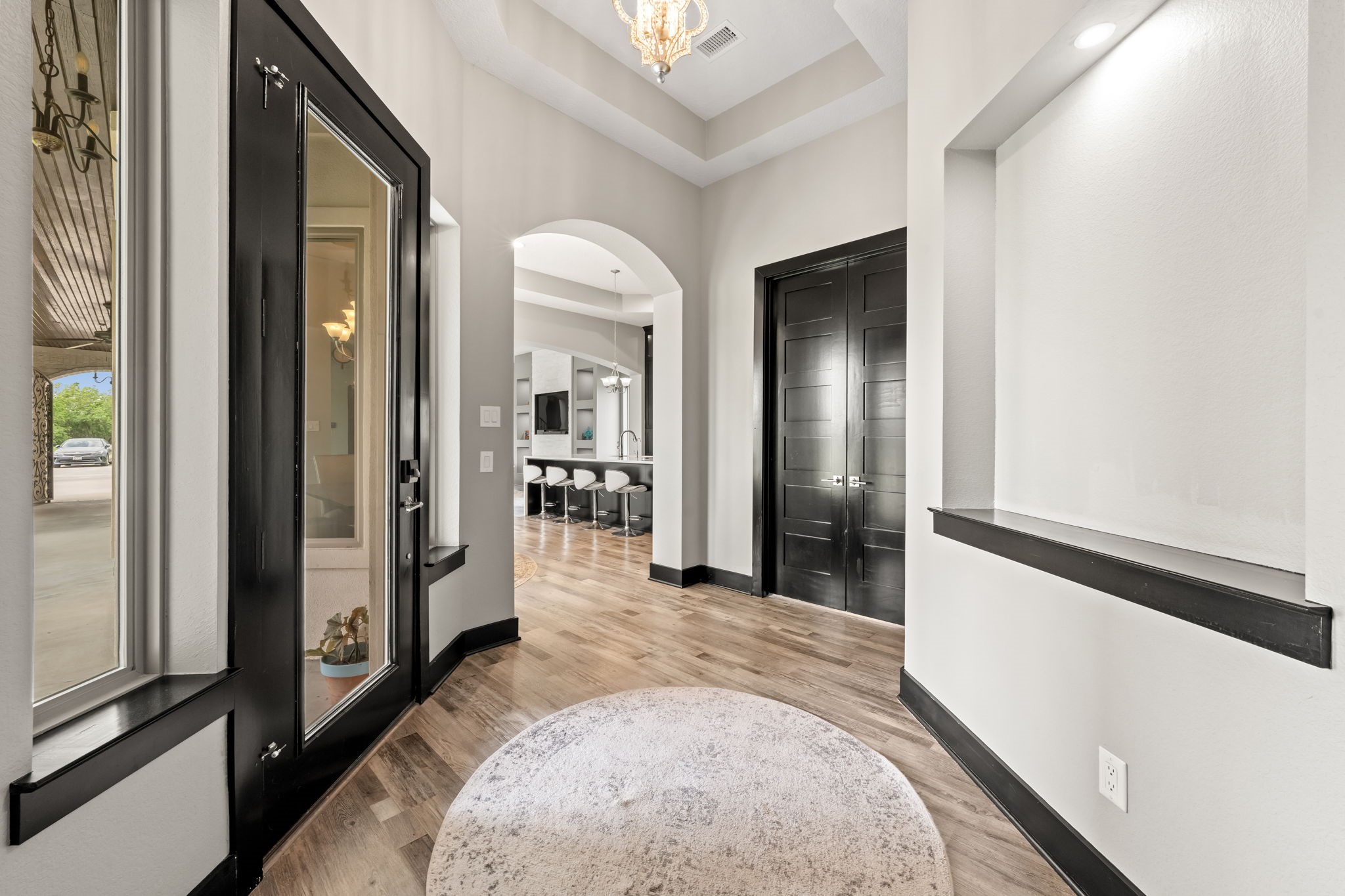 2856 Wolcek Road Crosby, TX 77532 - Photo 10 of 48 The elegant entryway features high ceilings, a stylish chandelier, and sleek engineered wood flooring. The black double doors and arched openings lead to a modern open-concept living area with a view of a spacious kitchen island, creating a welcoming and contemporary ambiance.