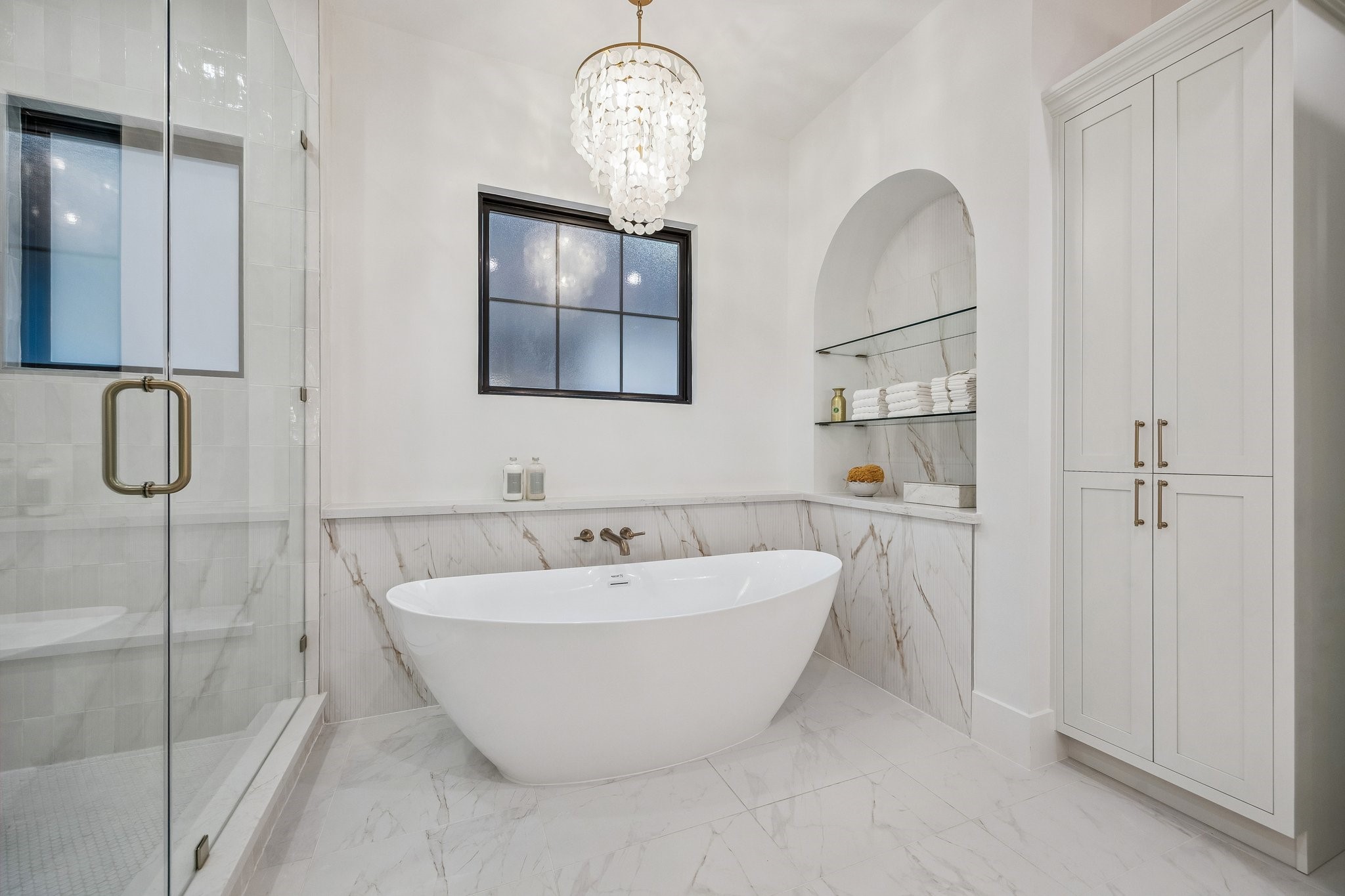 4034 Lanark Lane Houston, TX 77025 - Photo 21 of 32 Another view of the 71" Soaking Tub with a Wall-Mounted Tub Filler in Champagne Bronze and a Light Glossy Bronze Finish Tiered Chandelier with Natural White Shell. There is also open glass shelving in the alcove wall that holds neatly stacked towels, jars, and bath accessories. Linen Storage Closet for Toiletries and Towels.