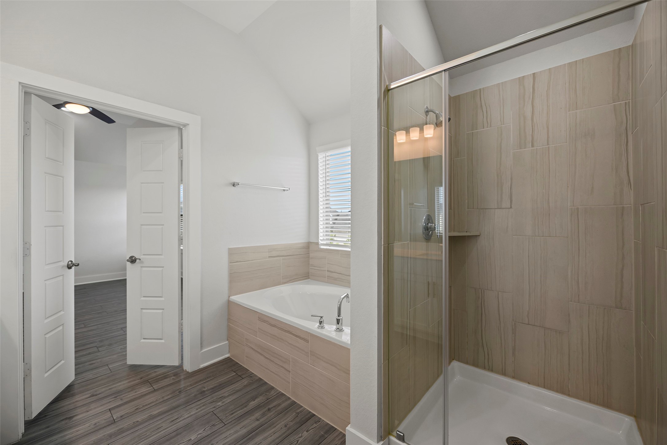 10603 Whitejacket Conroe, TX 77385 - Photo 18 of 42 Primary bathroom features a modern design with a spacious glass-enclosed shower, a large soaking tub, and sleek tile work. Natural light flows in through a window, enhancing the bright, open feel. Double doors lead to an adjacent room with wood-look flooring.