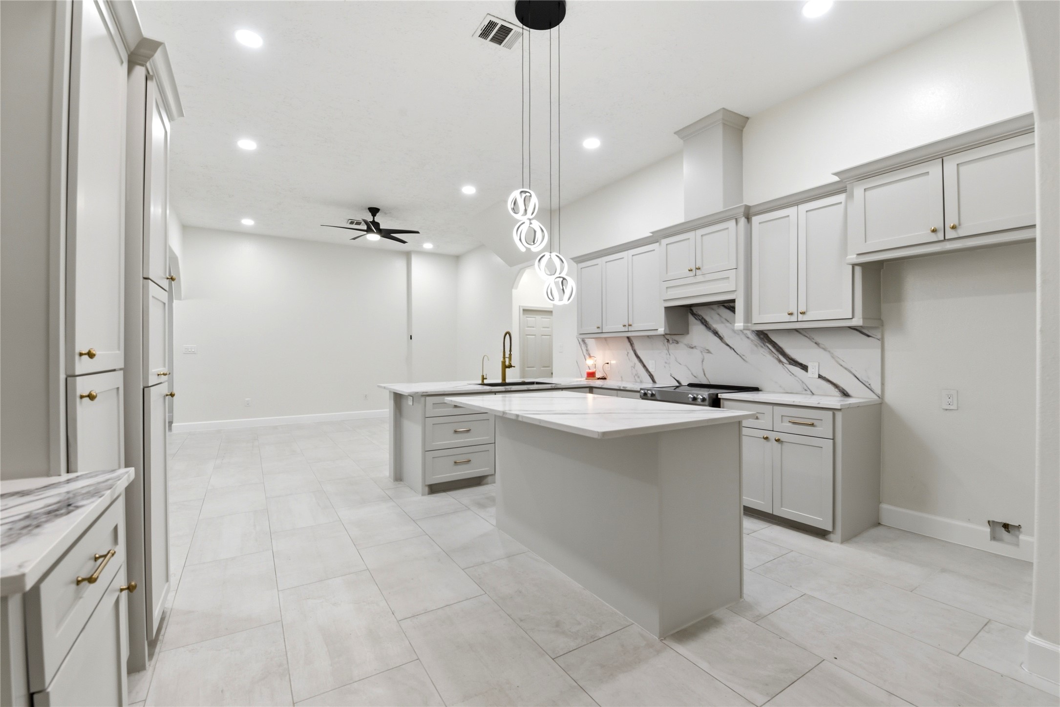 9403 Spaulding Street Houston, TX 77016 - Photo 12 of 50 This photo showcases a modern, spacious kitchen with sleek white cabinetry, a large island, marble backsplash, and contemporary lighting. The open layout flows into a bright living area with recessed lighting and a ceiling fan, creating an inviting and functional space.