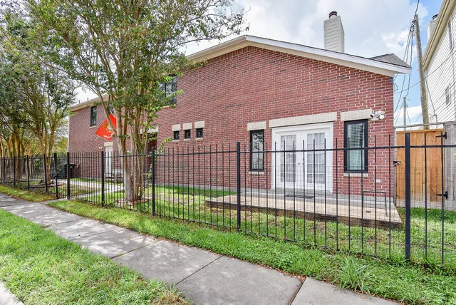 a view of a brick house in front of a yard with plants and large trees