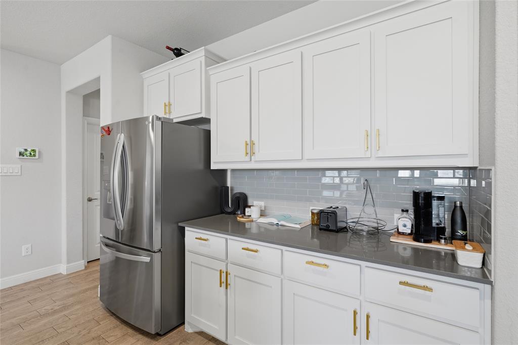1812 Ruffian Road Granbury, TX 76049 - Photo 13 of 37 White cabinetry with gold-tone hardware, dark countertops, a subway tile backsplash, stainless steel refrigerator, and wood-finish flooring