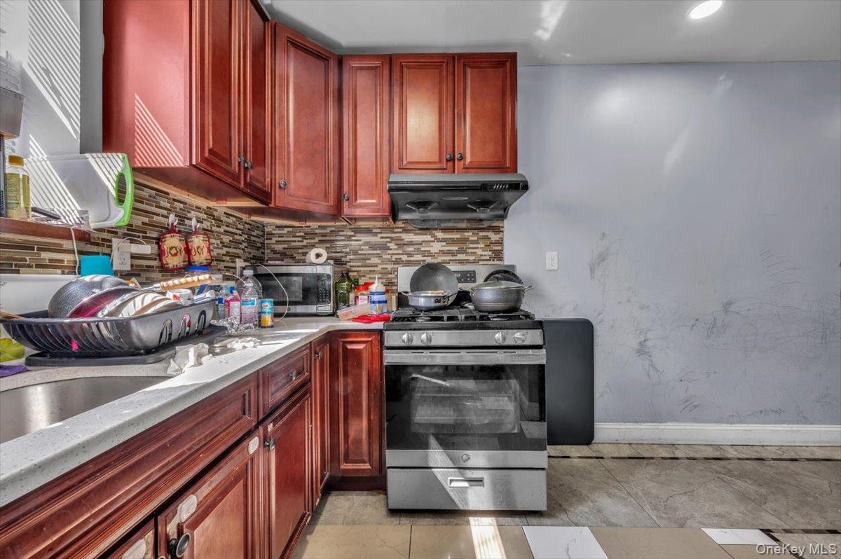 95-68 115th Street Queens, NY 11419 - Photo 19 of 28 Kitchen featuring appliances with stainless steel finishes, tasteful backsplash, reddish brown cabinets, light stone countertops, and light tile patterned floors