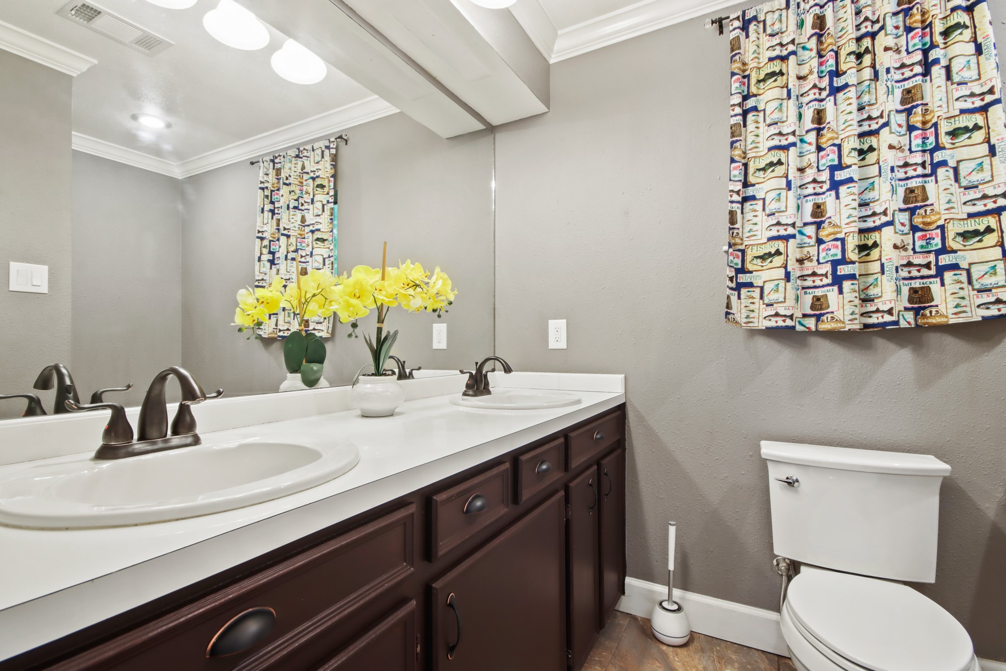 35 Susan Drive Cleveland, TX 77327 - Photo 20 of 33 This bathroom features a dual-sink vanity with dark wood cabinets, complemented by a large mirror. The neutral wall color enhances the space, and a patterned curtain adds a pop of color. Bright lighting and decorative flowers create a welcoming atmosphere.