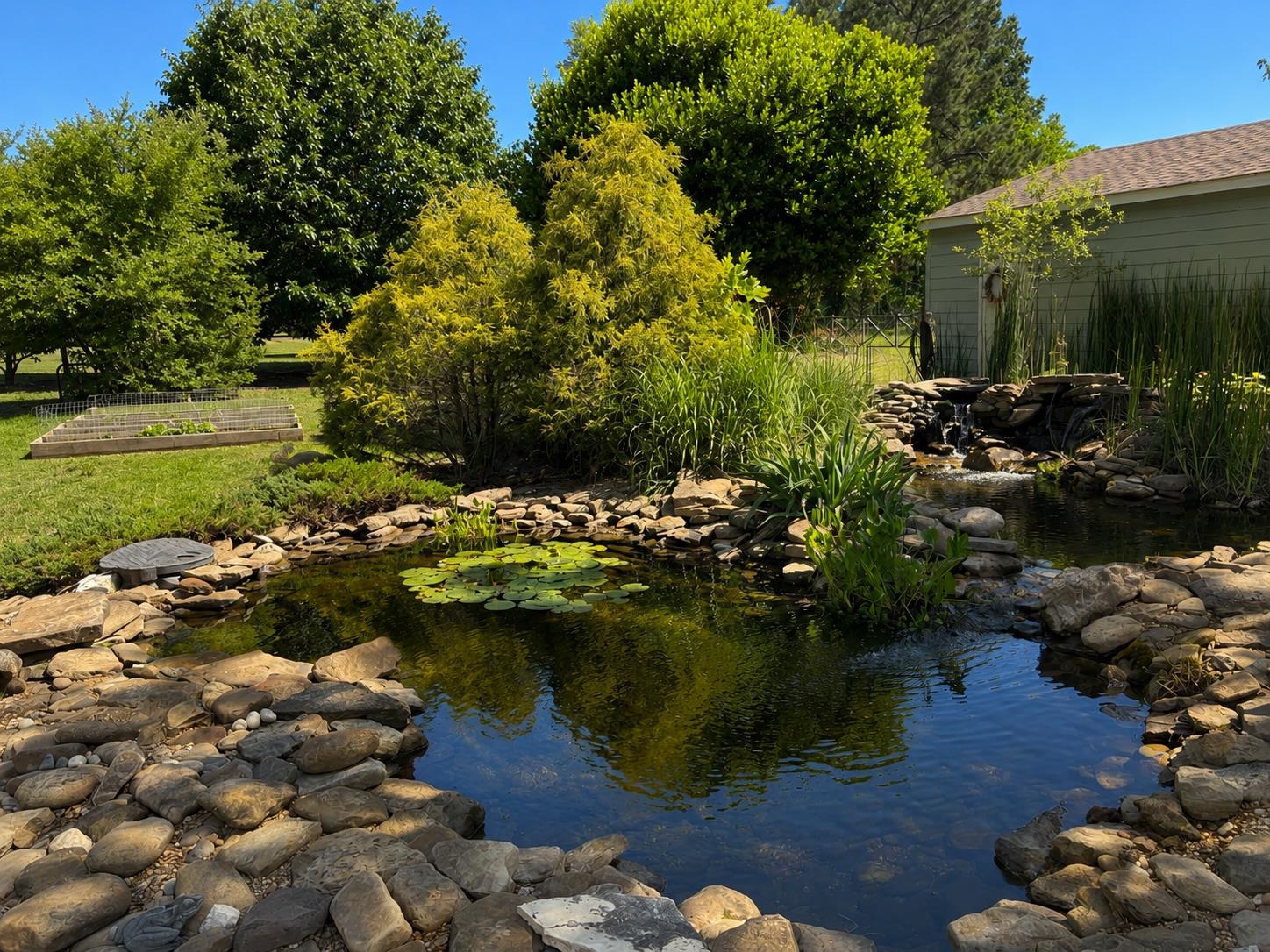 1946 Little John Road Millington, TN 38053 - Photo 16 of 20 Stone-lined pond with water feature, surrounded by lush greenery, mature trees, and a raised garden bed