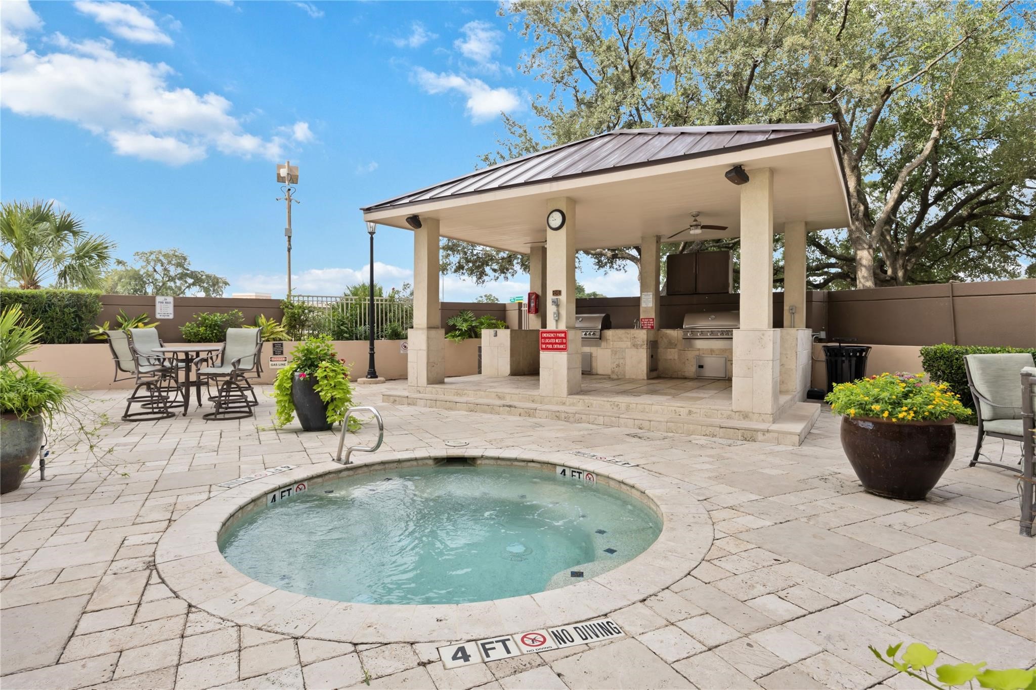 3525 Sage Road, Unit 1713 Houston, TX 77056 - Photo 23 of 25 This outdoor space features a cozy hot tub surrounded by a stylish stone deck. There's a covered grilling area with multiple barbecues and ample seating, ideal for entertaining. Lush greenery and a clear blue sky complete the inviting atmosphere.