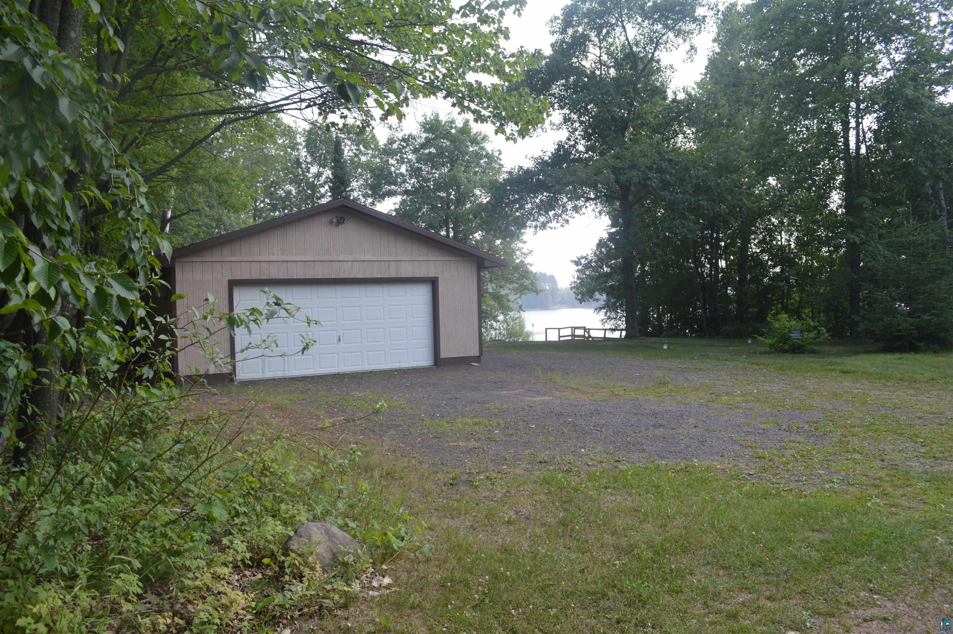 8467 County Rd P Lake Nebagamon, WI 54849 - Photo 2 of 25 Detached garage with a white overhead door, gravel driveway, and a lake visible through mature trees