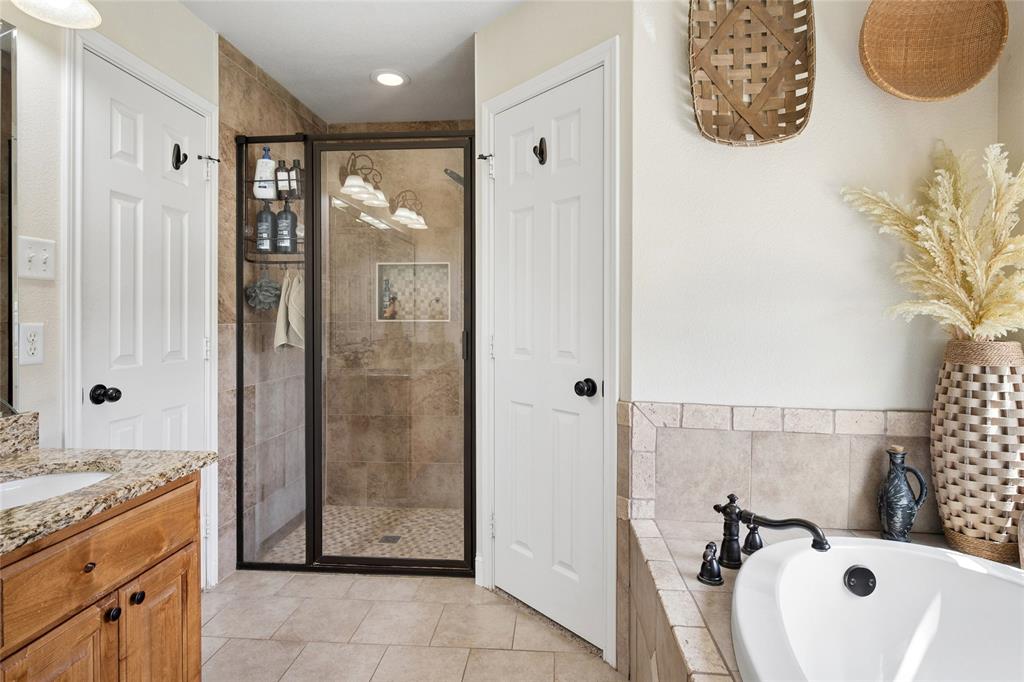 199 Newfield Lane Springtown, TX 76082 - Photo 20 of 38 The bathroom features a spacious walk-in shower with tiled walls and a glass enclosure, a white soaking tub with a dark-toned faucet, and a vanity with a granite countertop and wood cabinetry