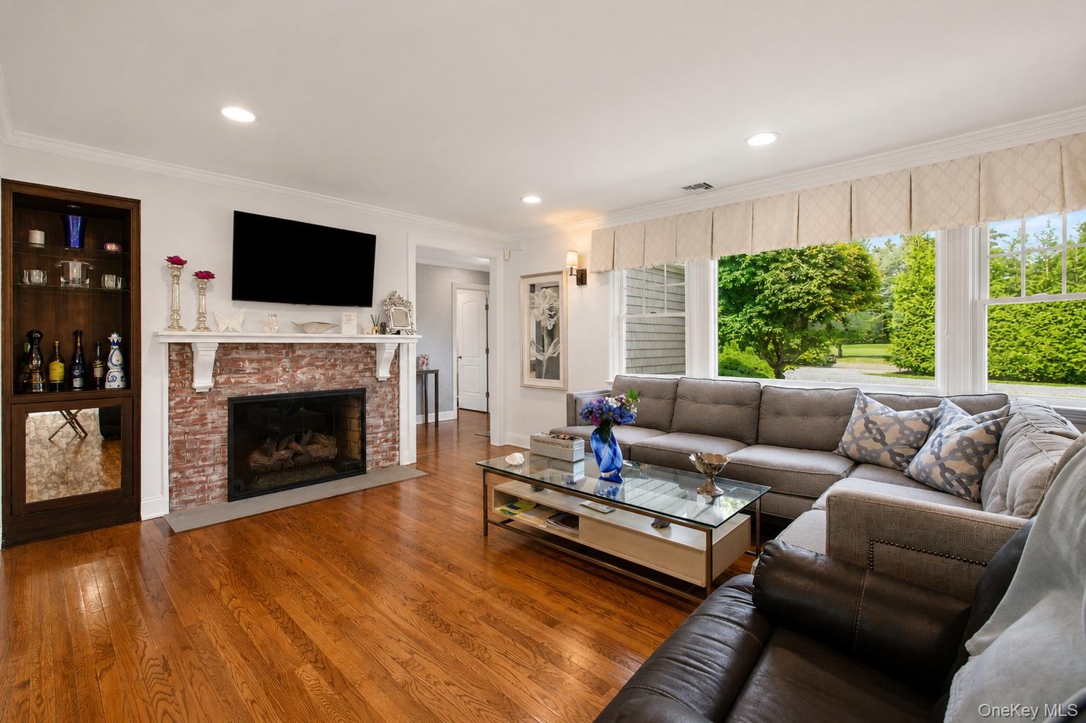 65 Shore Road Westhampton, NY 11977 - Photo 8 of 24 Living area featuring a red brick fireplace with a white mantel, recessed lighting, wood-finish flooring, a built-in display cabinet, and a large window arrangement