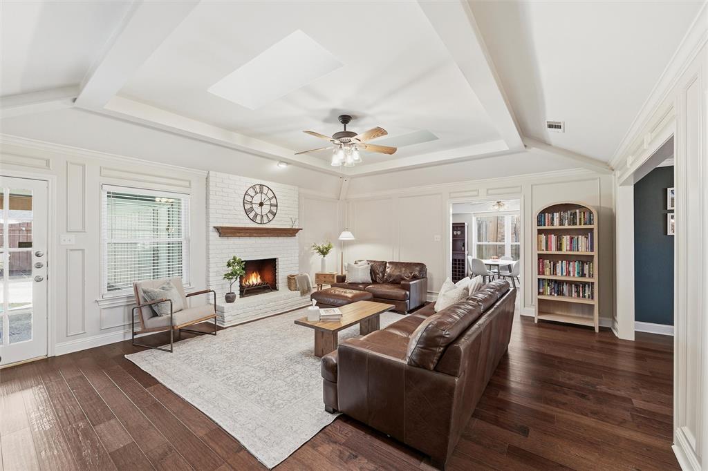 1704 Throwbridge Lane Plano, TX 75023 - Photo 11 of 40 Living room features tray ceiling with crown molding, skylights, and a ceiling fan with light kit. White-painted brick fireplace includes a raised hearth, wood mantel, and gas starter, with wide cased openings and glass-panel exterior door connecting the space to adjacent areas. Hardwood flooring extends throughout the room.