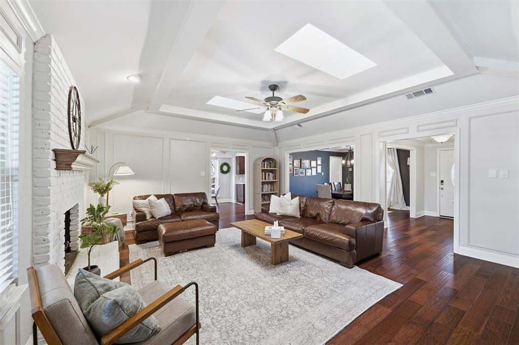 1704 Throwbridge Lane Plano, TX 75023 - Photo 12 of 40 Living room features tray ceiling with crown molding, skylights, and a ceiling fan with light kit. White-painted brick fireplace includes a raised hearth, wood mantel, and gas starter, with wide cased openings and glass-panel exterior door connecting the space to adjacent areas. Hardwood flooring extends throughout the room.