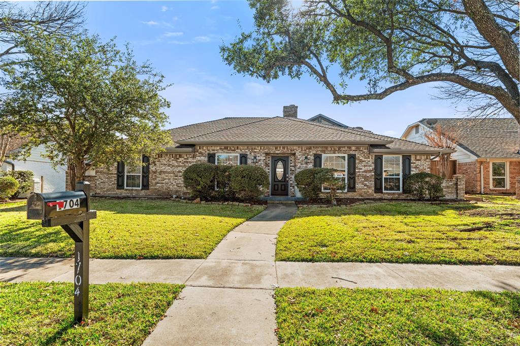 1704 Throwbridge Lane Plano, TX 75023 - Photo 4 of 40 Front exterior features traditional brick construction, multiple front-facing windows with decorative shutters, and a centered front entry. A straight concrete walkway leads from the sidewalk to the front door, bordered by maintained lawn, trimmed shrubs, and mature trees providing natural shade.