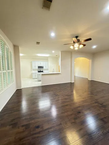 an empty room with wooden floor a ceiling fan and kitchen view