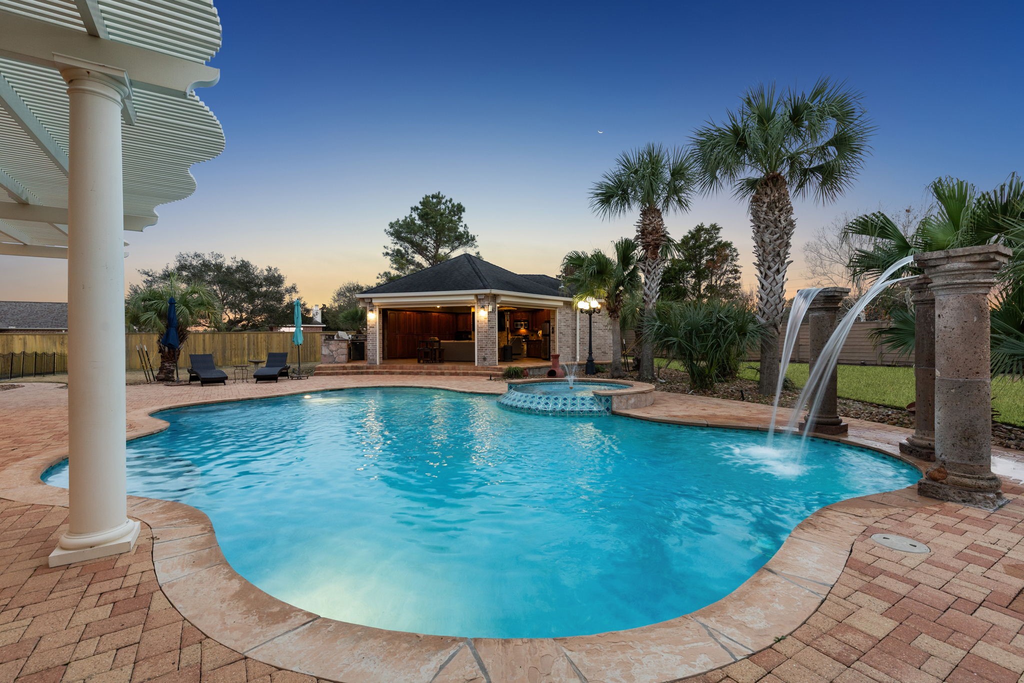 2202 Britton Ridge Drive Katy, TX 77494 - Photo 3 of 41 An entertainer’s dream at sunset—this resort-style pool and spa features elegant water features, expansive paver decking, and a fully equipped summer kitchen/cabana with fireplace, creating a private outdoor oasis perfect for upscale gatherings and relaxed evenings at home.