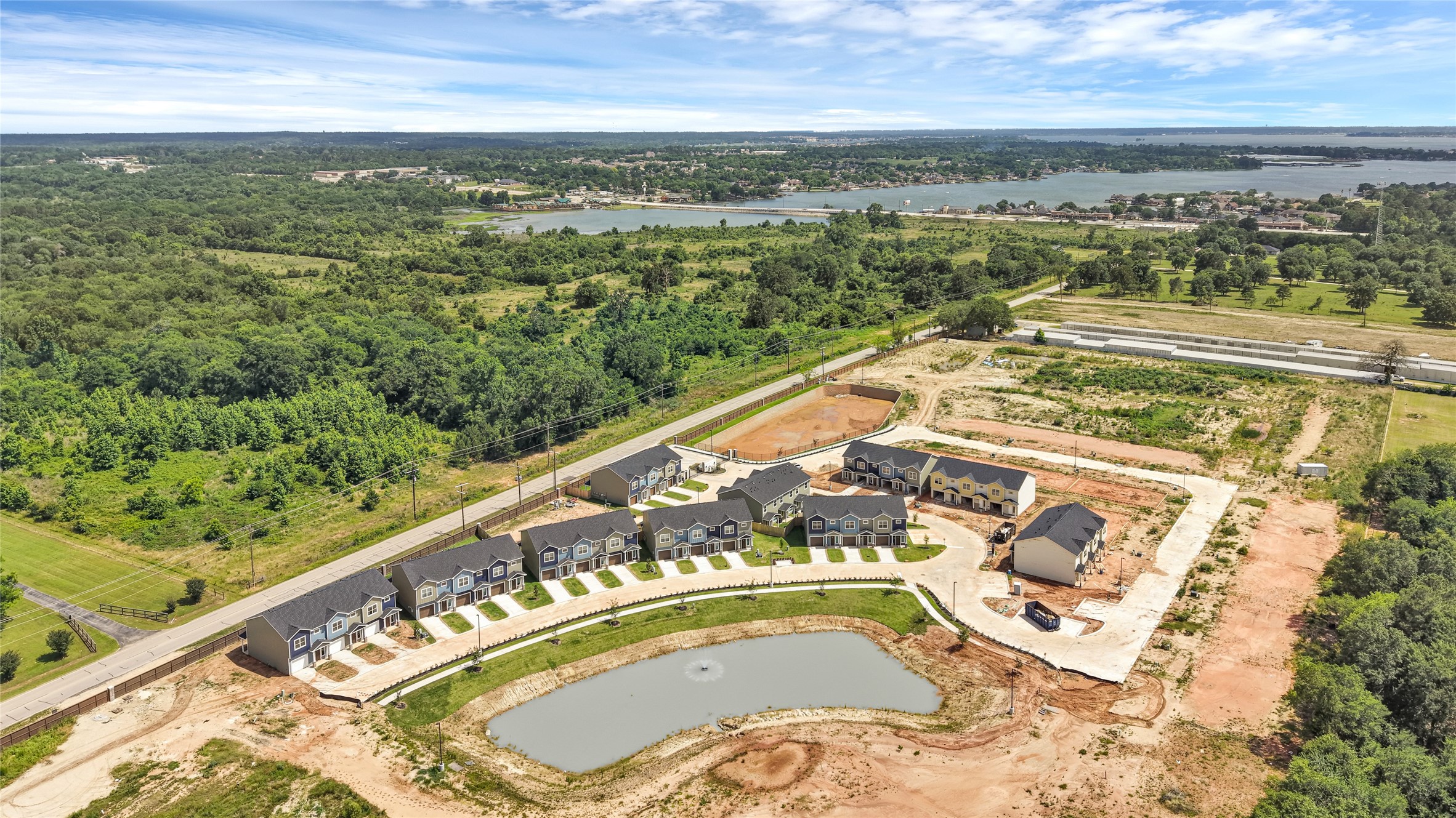 14010 Longstreet Road, Unit 2 Willis, TX 77318 - Photo 29 of 29 This photo shows a new residential development with rows of modern townhomes, neatly arranged around a central pond. The community is surrounded by lush greenery and has a picturesque view of a nearby lake, offering a serene and attractive setting.