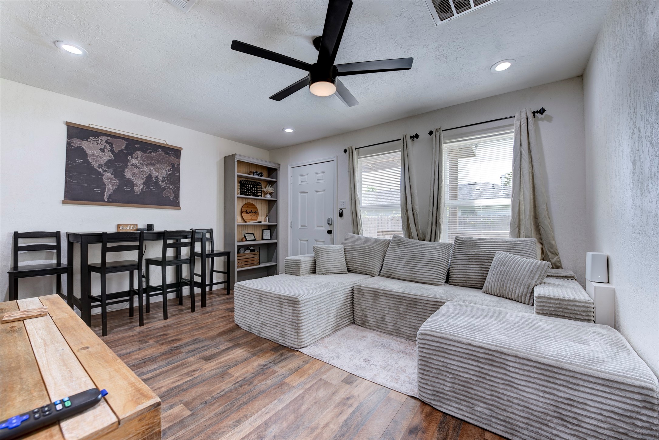1121 Panama Street Houston, TX 77009 - Photo 6 of 35 Living and dining area featuring dark wood-look LVP flooring, oversized black ceiling fan with light kit, recessed lighting, dual oversized windows with grommet linen curtain panels on black rod hardware, five-shelf built-in bookcase with beadboard back panel, six-panel entry door with smart lock, and black bar-height dining set.