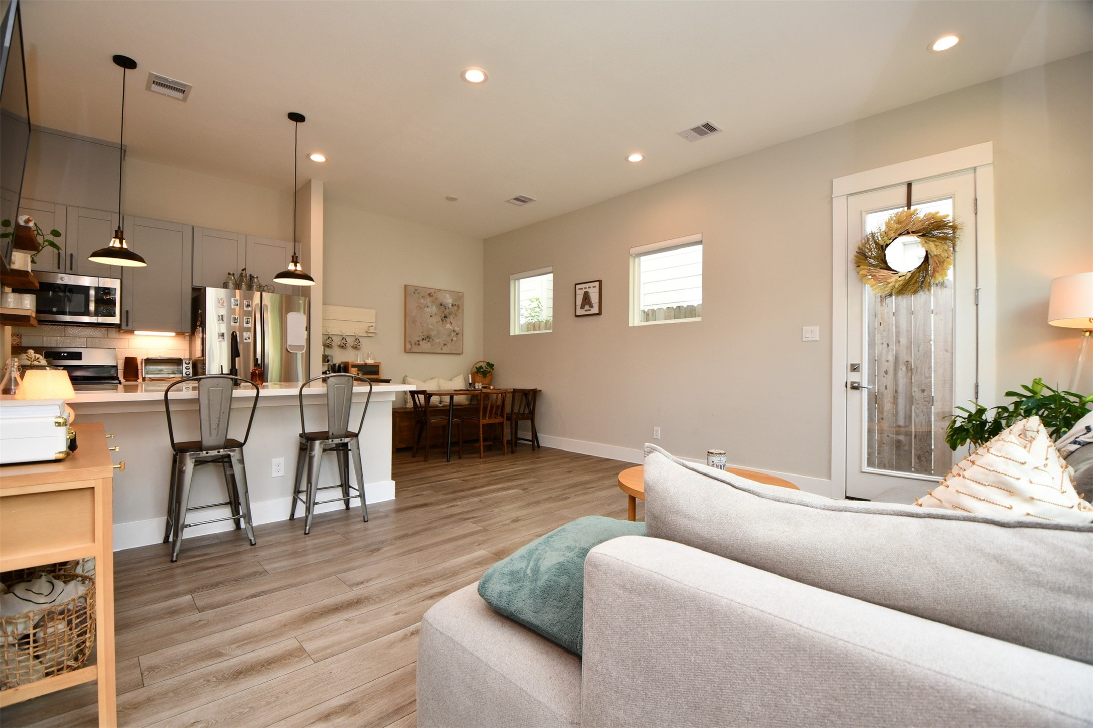 1239 West Tidwell Road Houston, TX 77091 - Photo 12 of 35 This photo showcases a modern, open-concept living area with sleek hardwood floors and ample natural light. The kitchen features contemporary appliances and a breakfast bar with pendant lighting, while the adjacent dining space offers a cozy setting. A glass door with a decorative wreath leads to an outdoor area, enhancing the inviting atmosphere.