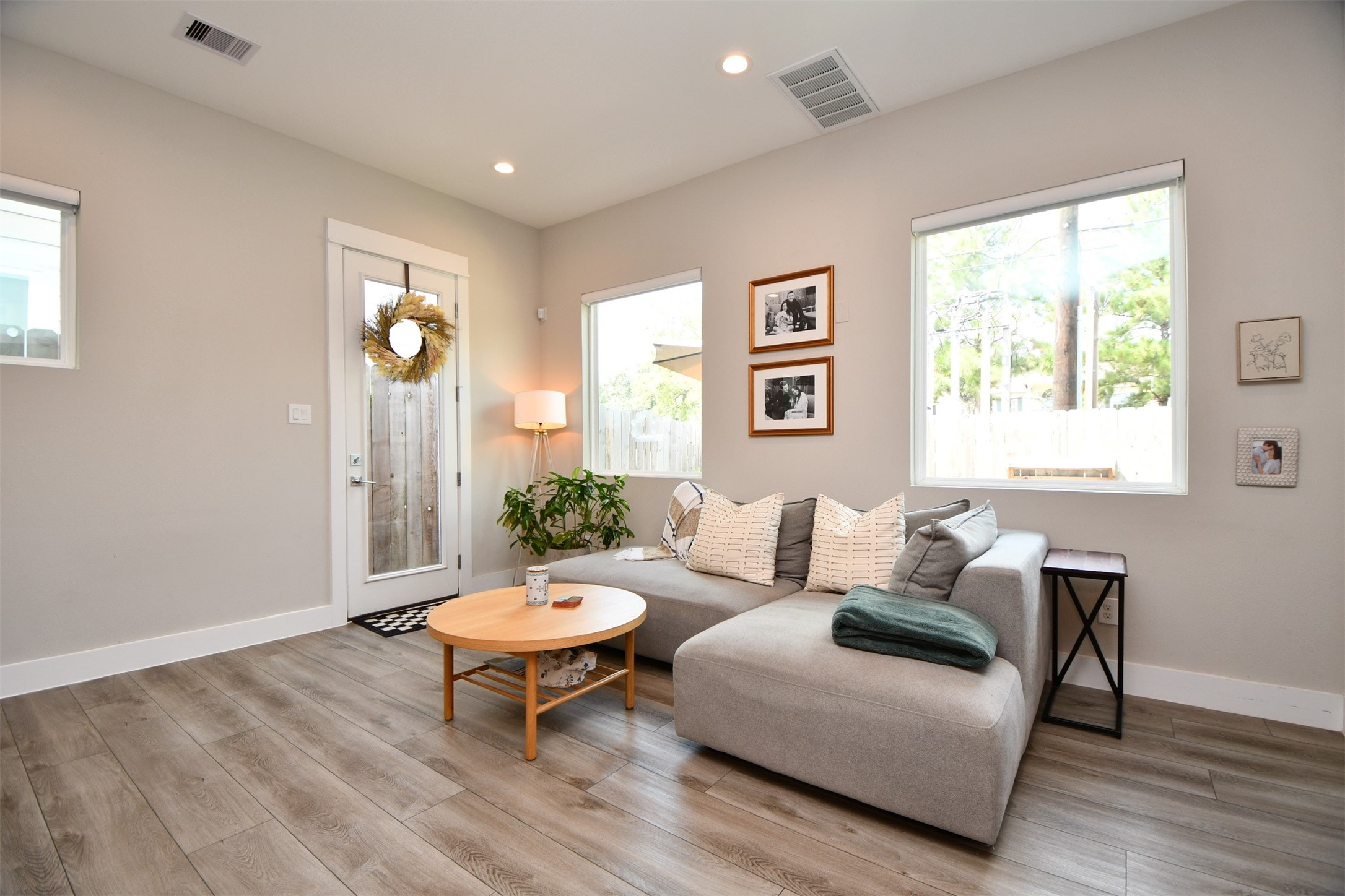 1239 West Tidwell Road Houston, TX 77091 - Photo 14 of 35 Bright, cozy living area with modern wood flooring, a comfortable gray sectional sofa, and large windows providing natural light. Features a stylish round coffee table, wall art, and a decorative wreath on the door. Perfect for relaxation and entertaining.
