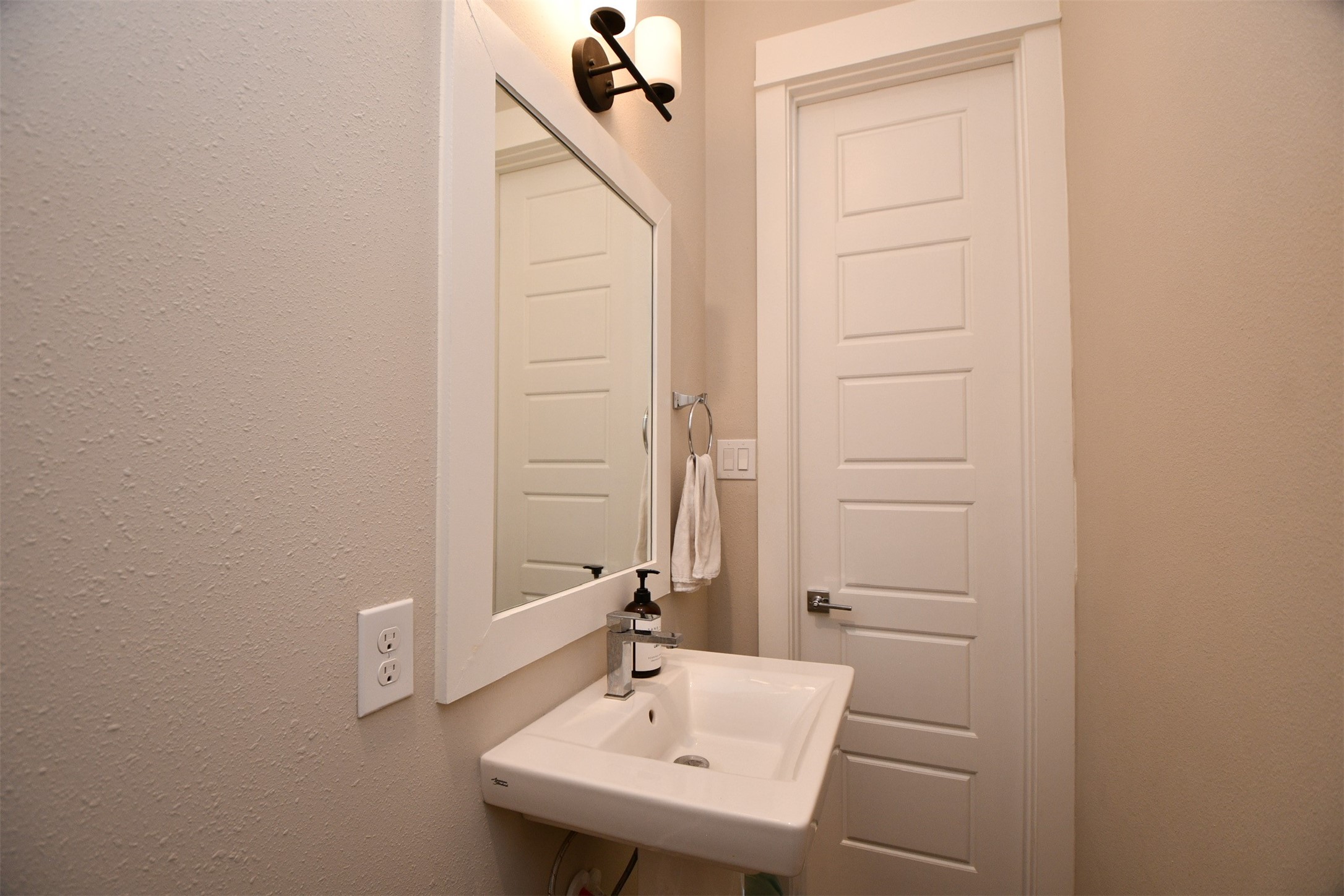 1239 West Tidwell Road Houston, TX 77091 - Photo 15 of 35 This photo features a compact, modern bathroom with a sleek white sink, a large mirror, and an elegant light fixture above. The neutral wall color complements the white paneled door, creating a clean and inviting space.