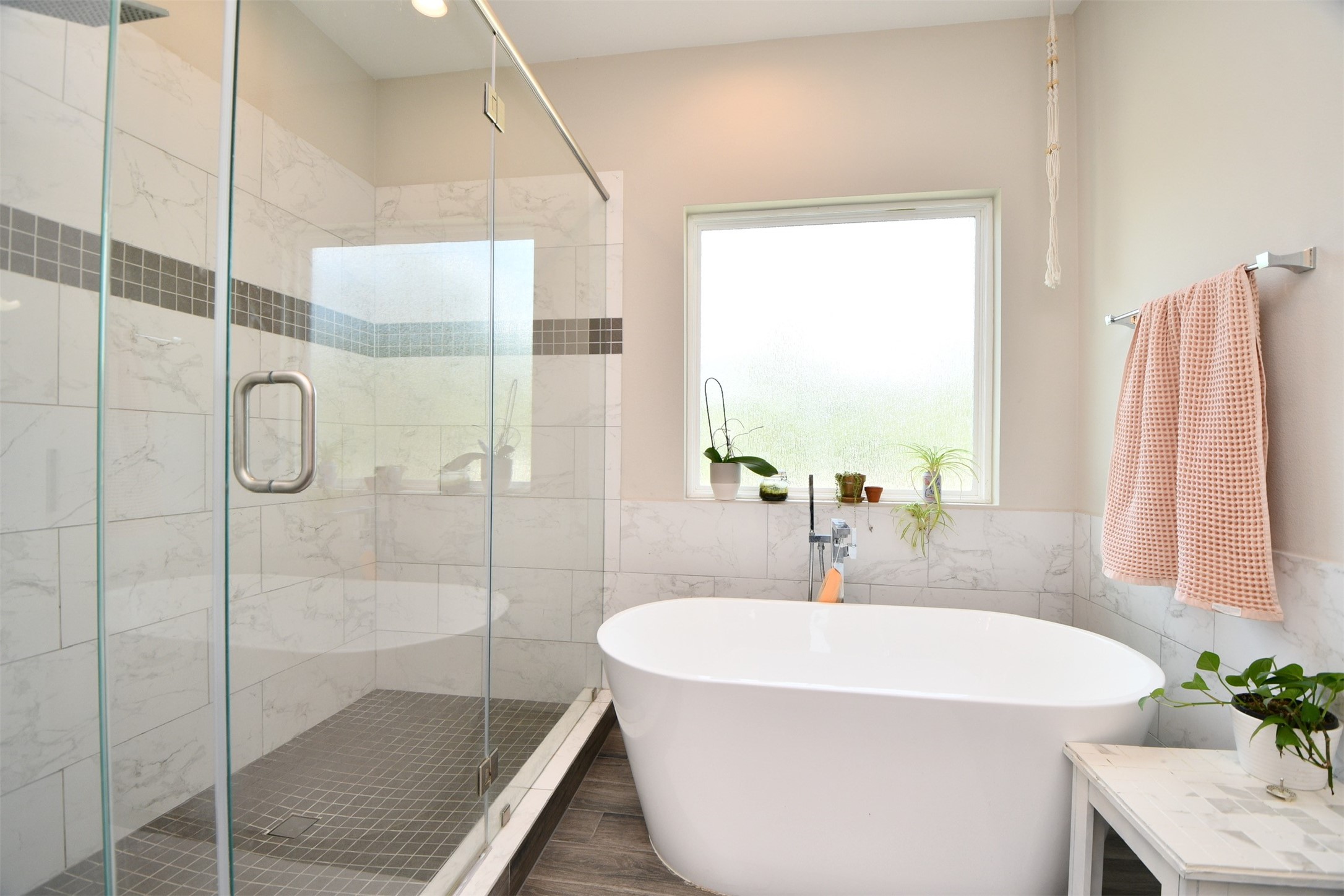 1239 West Tidwell Road Houston, TX 77091 - Photo 23 of 35 This modern bathroom features a spacious glass-enclosed shower with elegant tile work and a sleek, freestanding bathtub. Natural light pours in through a large frosted window, creating a bright and inviting atmosphere. The space is accented with plants and soft peach towels, adding a touch of warmth and style.