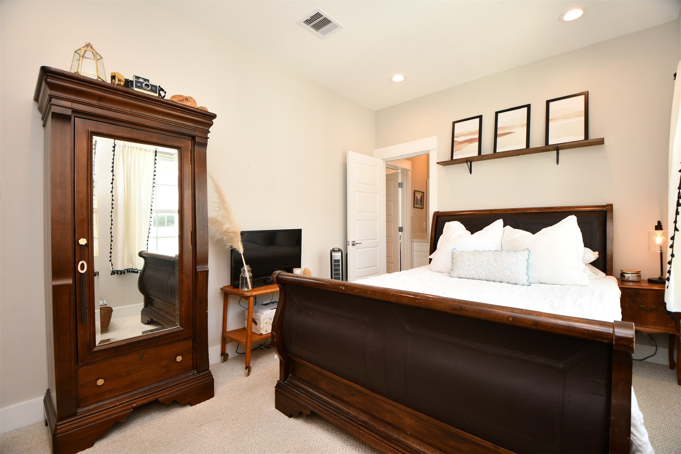 1239 West Tidwell Road Houston, TX 77091 - Photo 26 of 35 This cozy bedroom features a stylish dark wood bed and matching wardrobe with a mirrored door. The room is well-lit with recessed lighting and natural light from a window. A small shelf with framed art adds a decorative touch above the bed. Light walls and carpet create an inviting atmosphere.