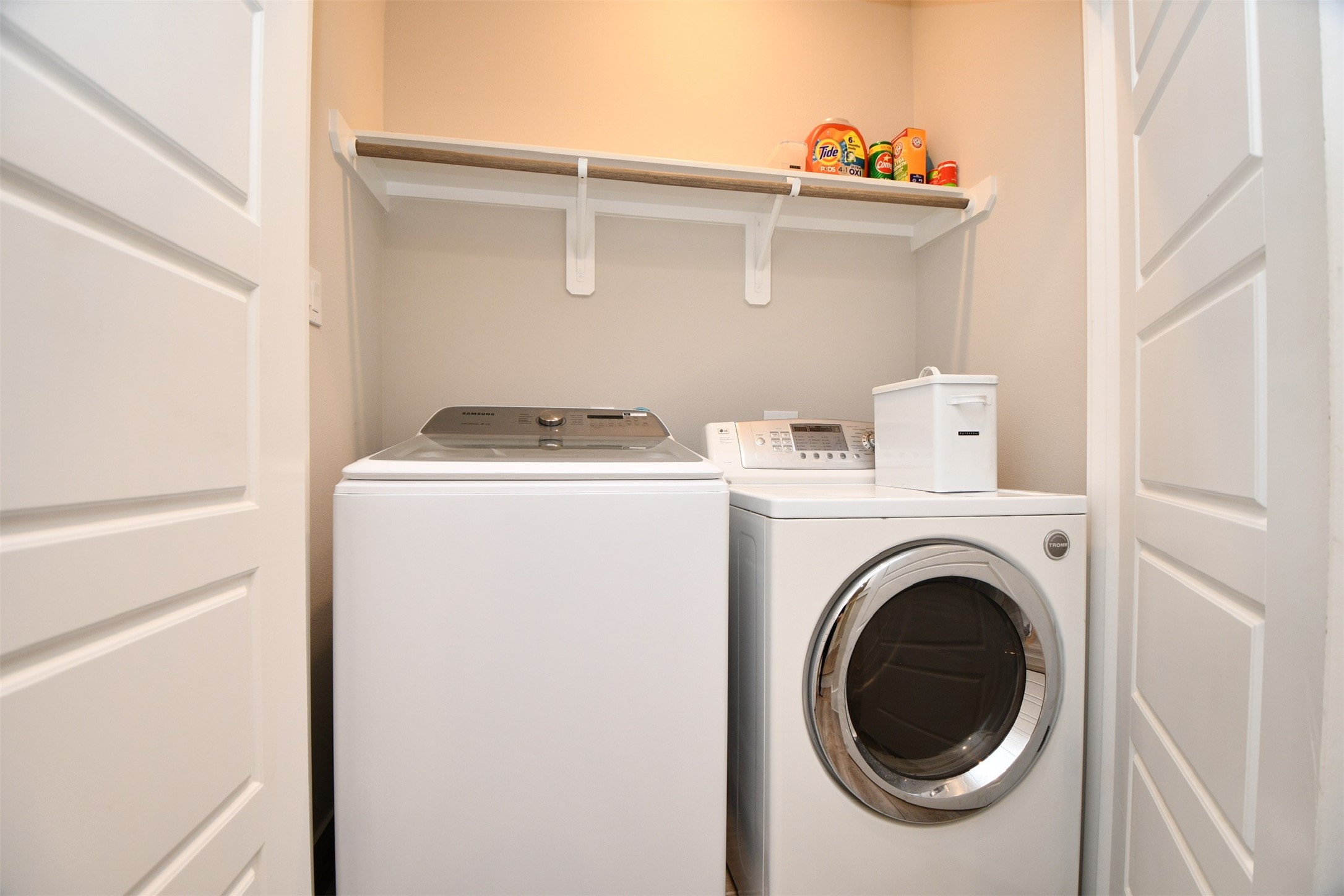 1239 West Tidwell Road Houston, TX 77091 - Photo 27 of 35 This photo shows a compact laundry closet with a modern washer and dryer set. Above the appliances, there's a practical shelf for storage. The space is well-lit and efficiently designed for convenience.