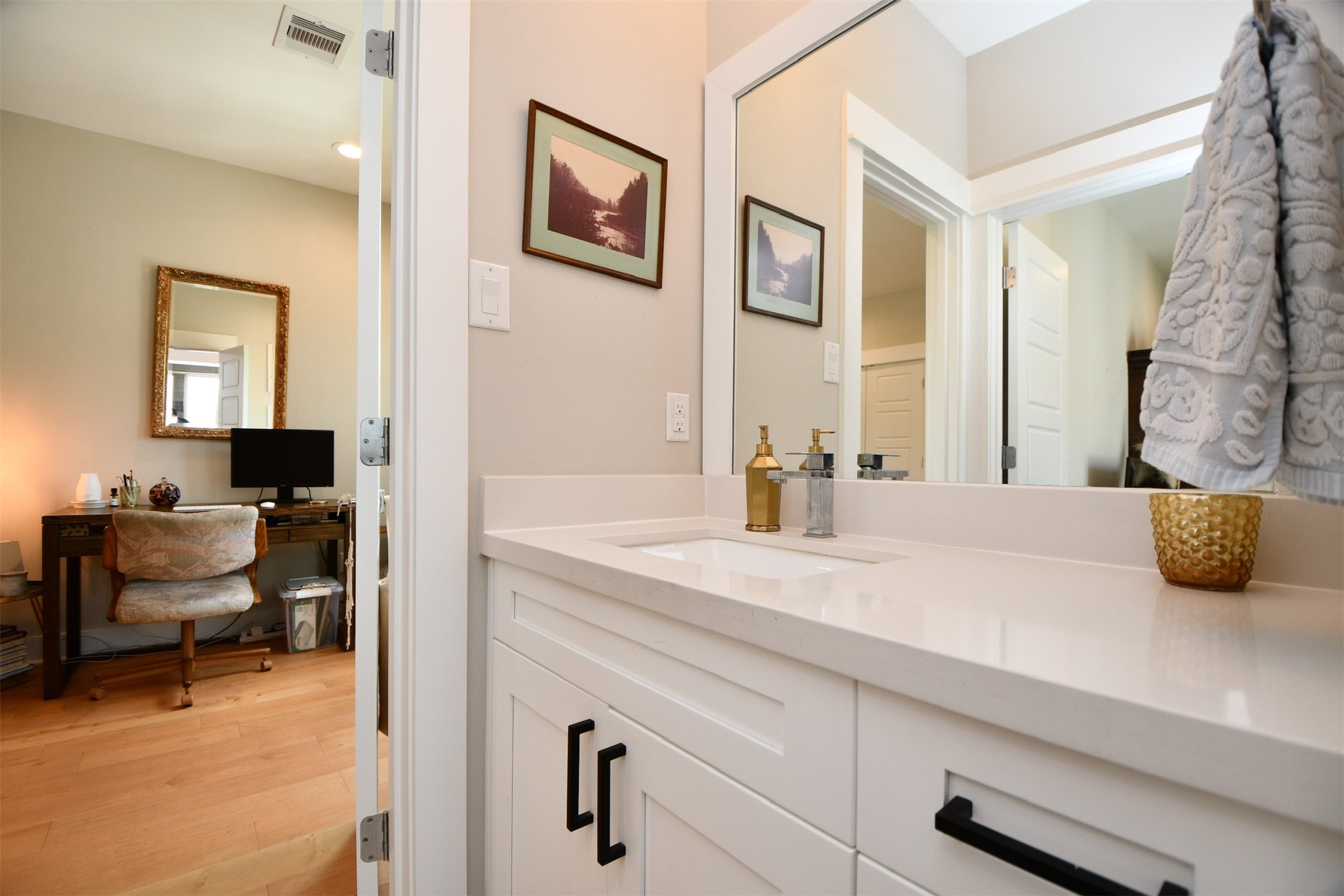 1239 West Tidwell Road Houston, TX 77091 - Photo 29 of 35 This photo shows a modern bathroom with a sleek white vanity featuring a large mirror and decorative items. It opens into a cozy room with a desk, chair, and wall art, offering a seamless connection between spaces.